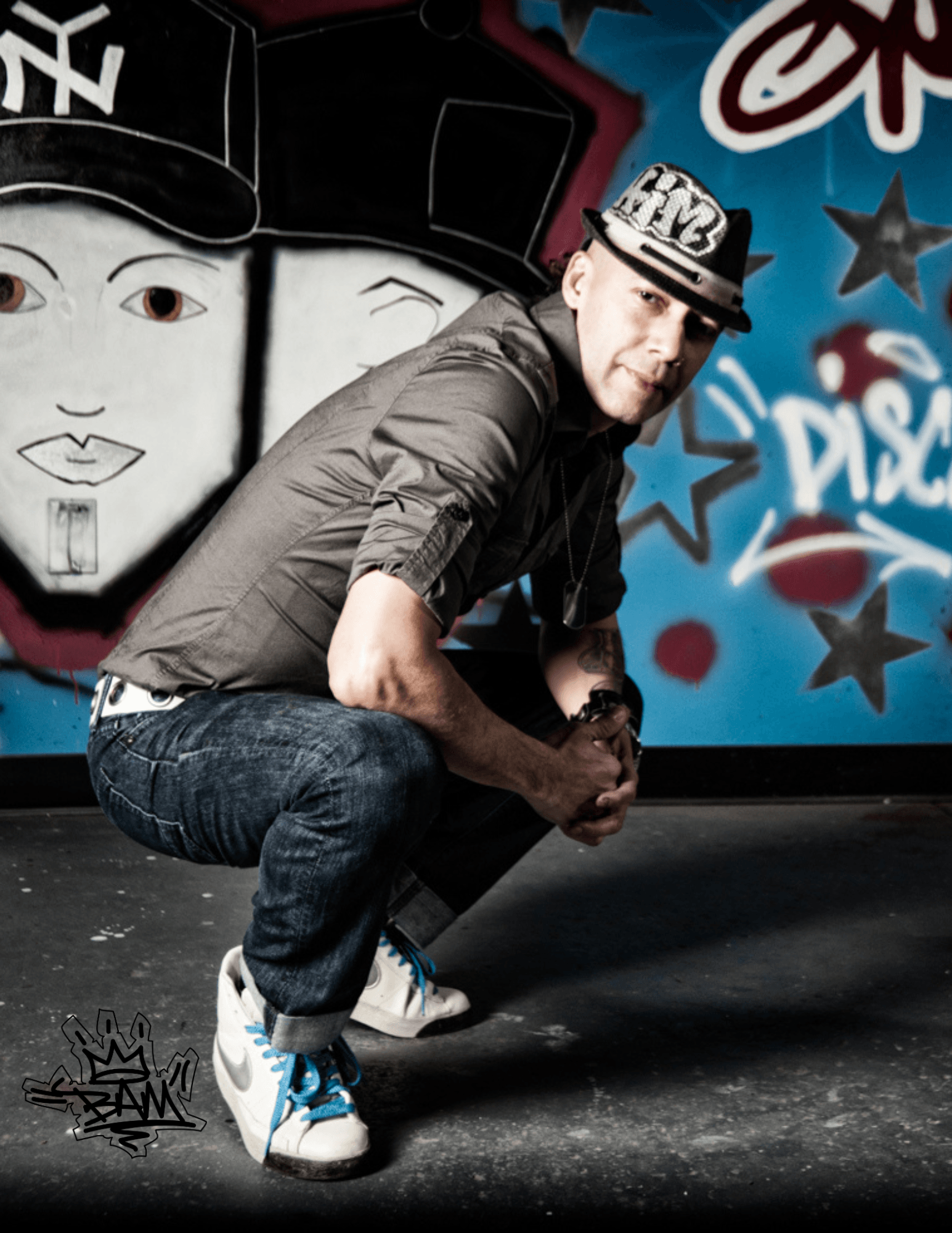 Man crouching in front of graffiti wall with faces and stars, wearing a cap, gray shirt, and jeans.