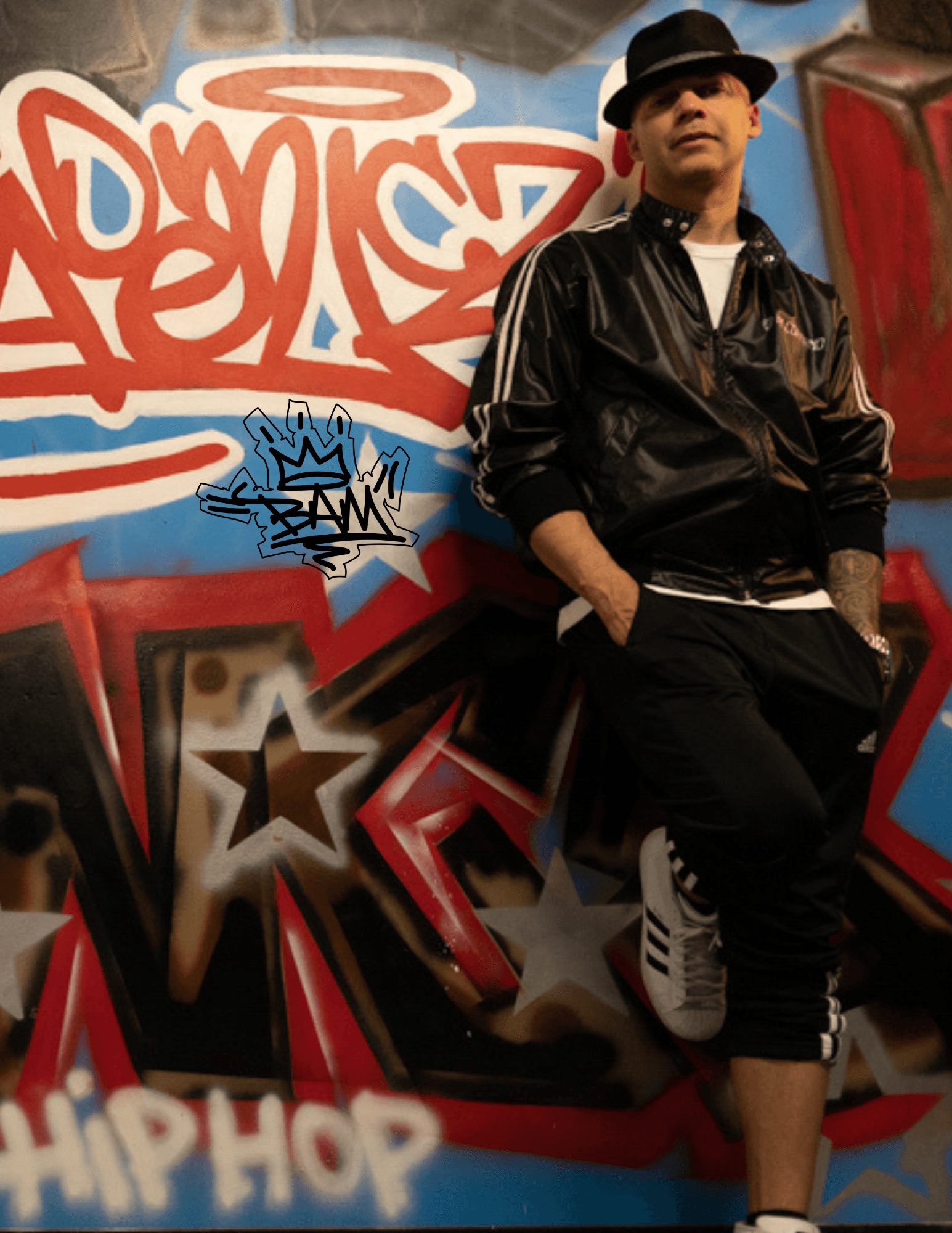 Man in black jacket and hat leaning against graffiti wall with stars and hip hop text.