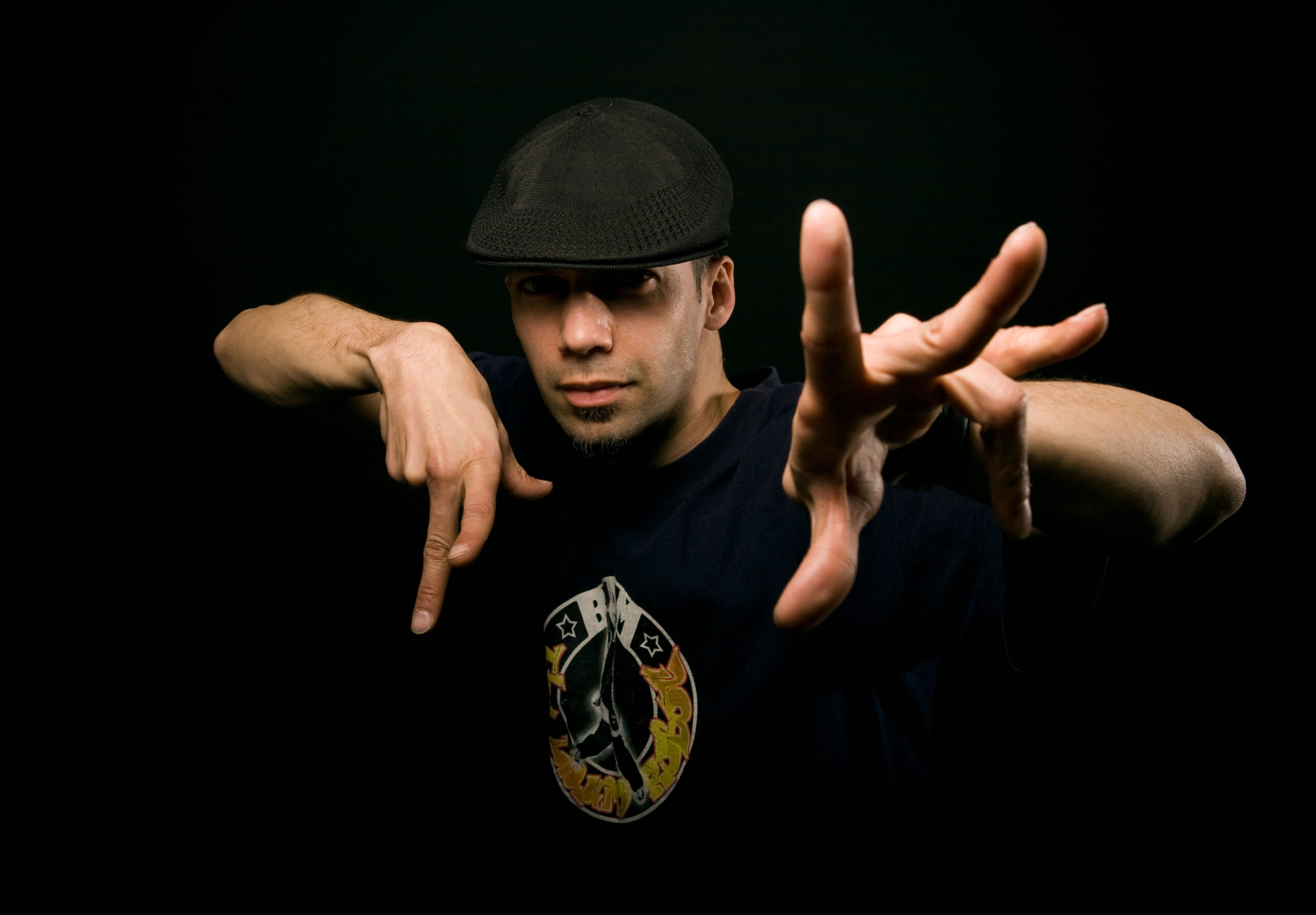 Person wearing a black cap and shirt, making a gesture with hands against a dark background.