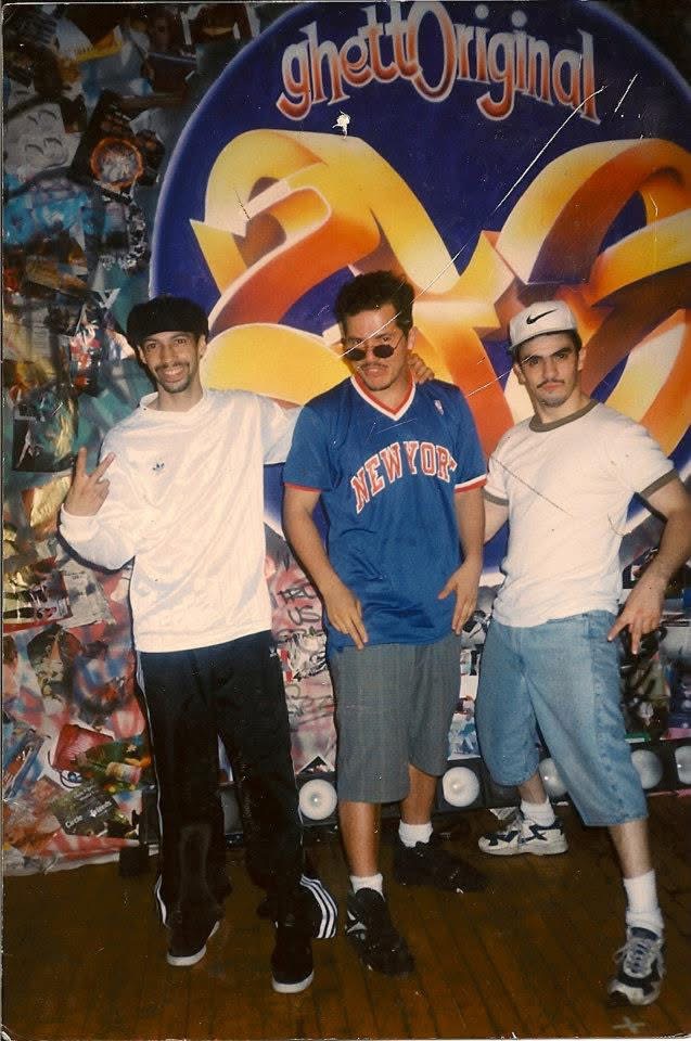 Three young men stand in front of a colorful backdrop with the text 'Ghetto Original' and a large yellow ribbon design.