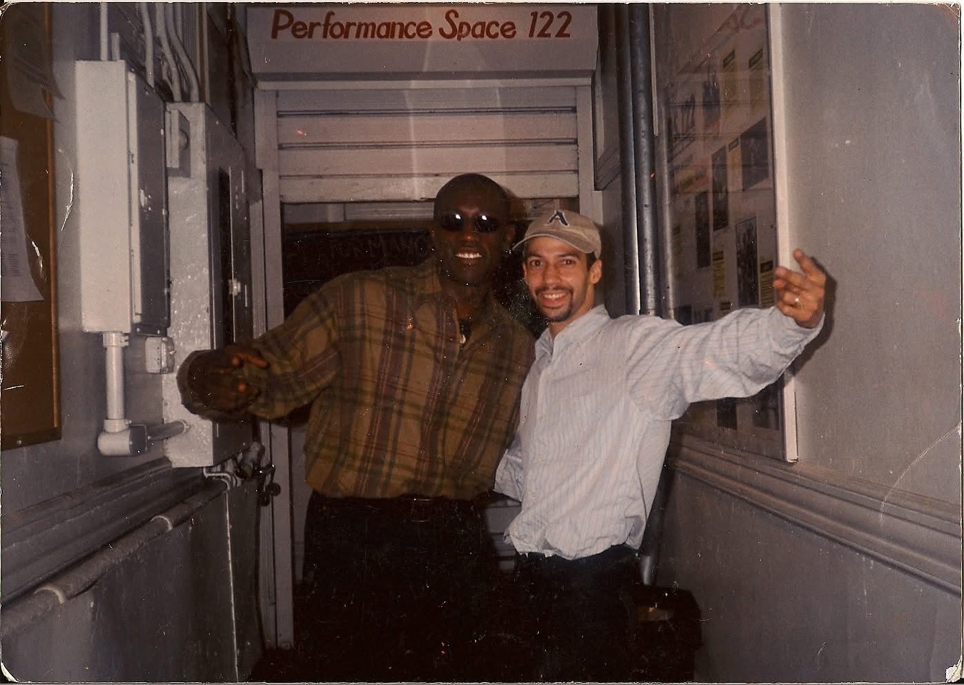Two men standing in a narrow hallway, smiling and posing with arms around each other, behind a sign that reads 'Performance Space 122'.