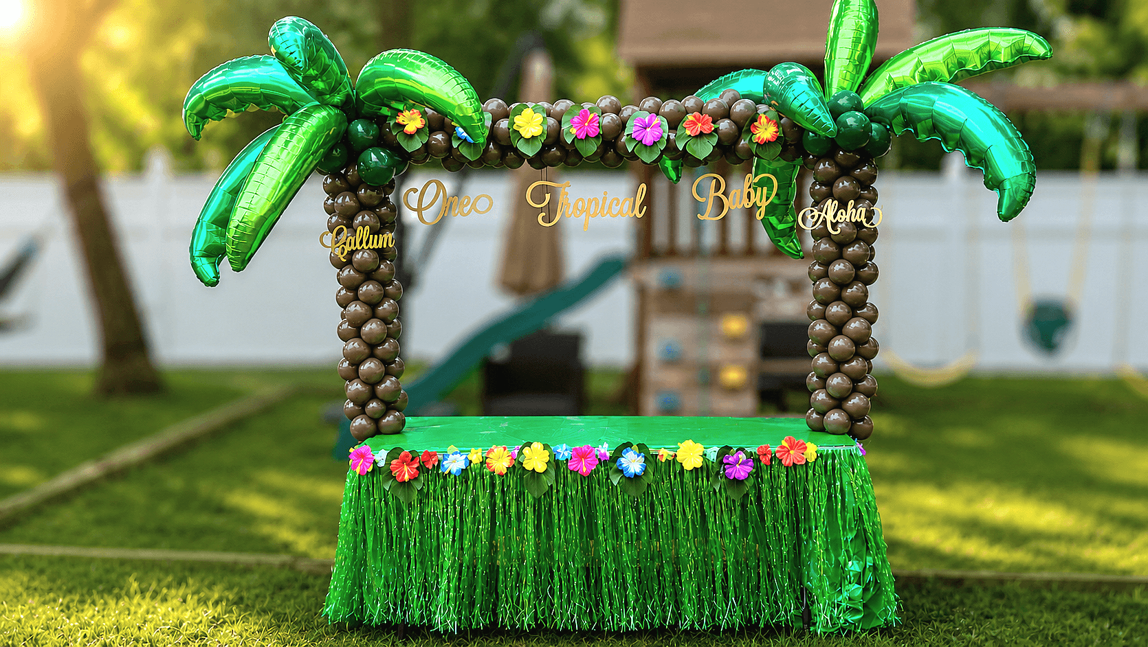 Colorful balloon tiki bar with palm trees, flowers, and green fringe, set outdoors on grass with trees and a fence in background.