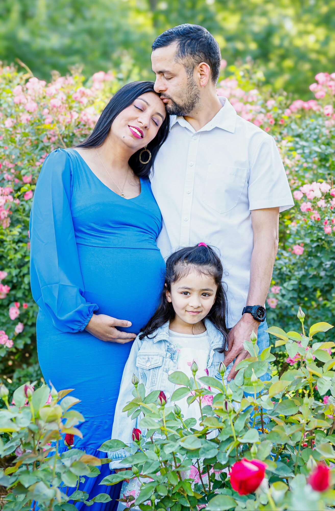 Pregnant woman in blue dress, man, and young girl outdoors with pink flowers and greenery.