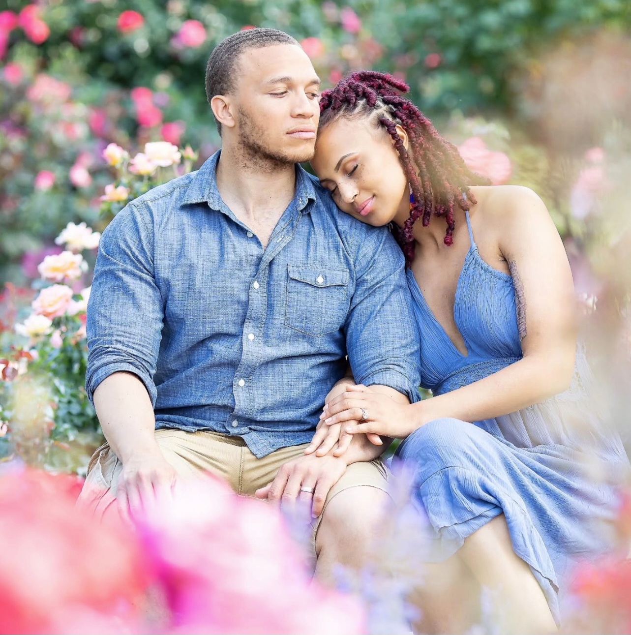 A couple sitting close together in a garden with colorful flowers, the woman resting her head on the man's shoulder.