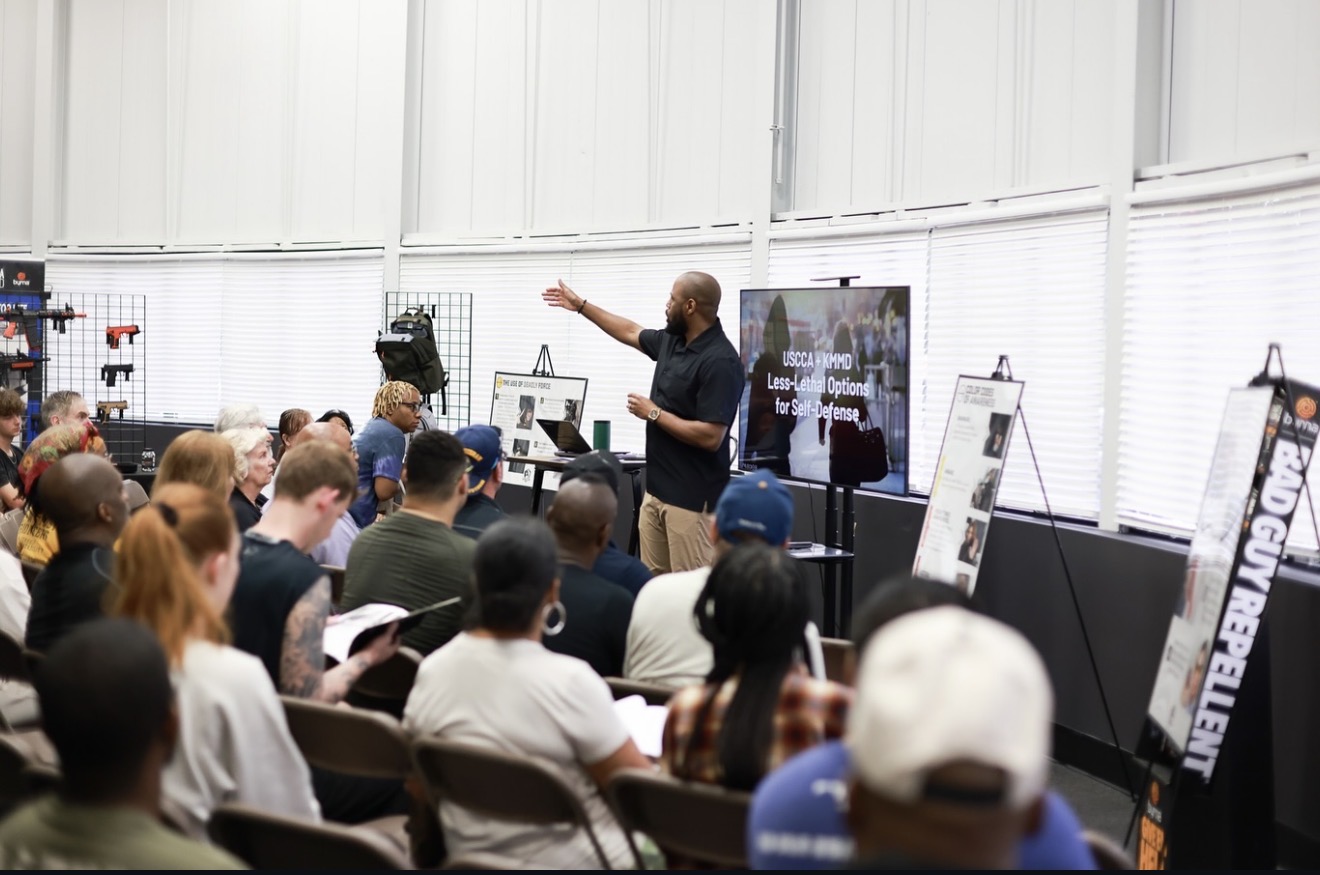 A speaker stands in front of a group, gesturing with one hand, with presentation screens and posters behind him.