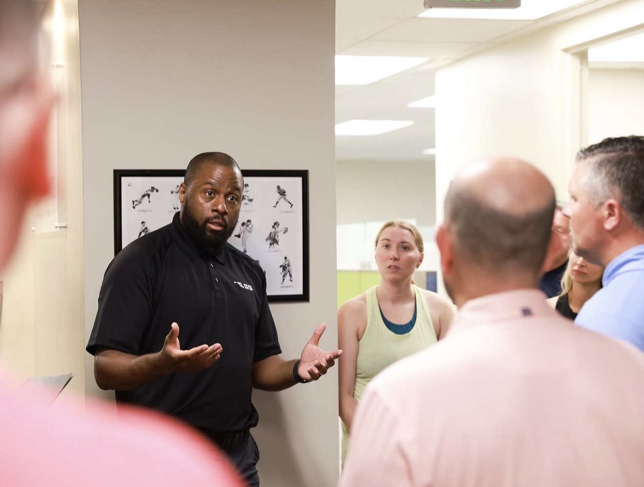 Man in black shirt speaking to a group of people in a room with white walls and a framed picture, woman in green tank top listening.