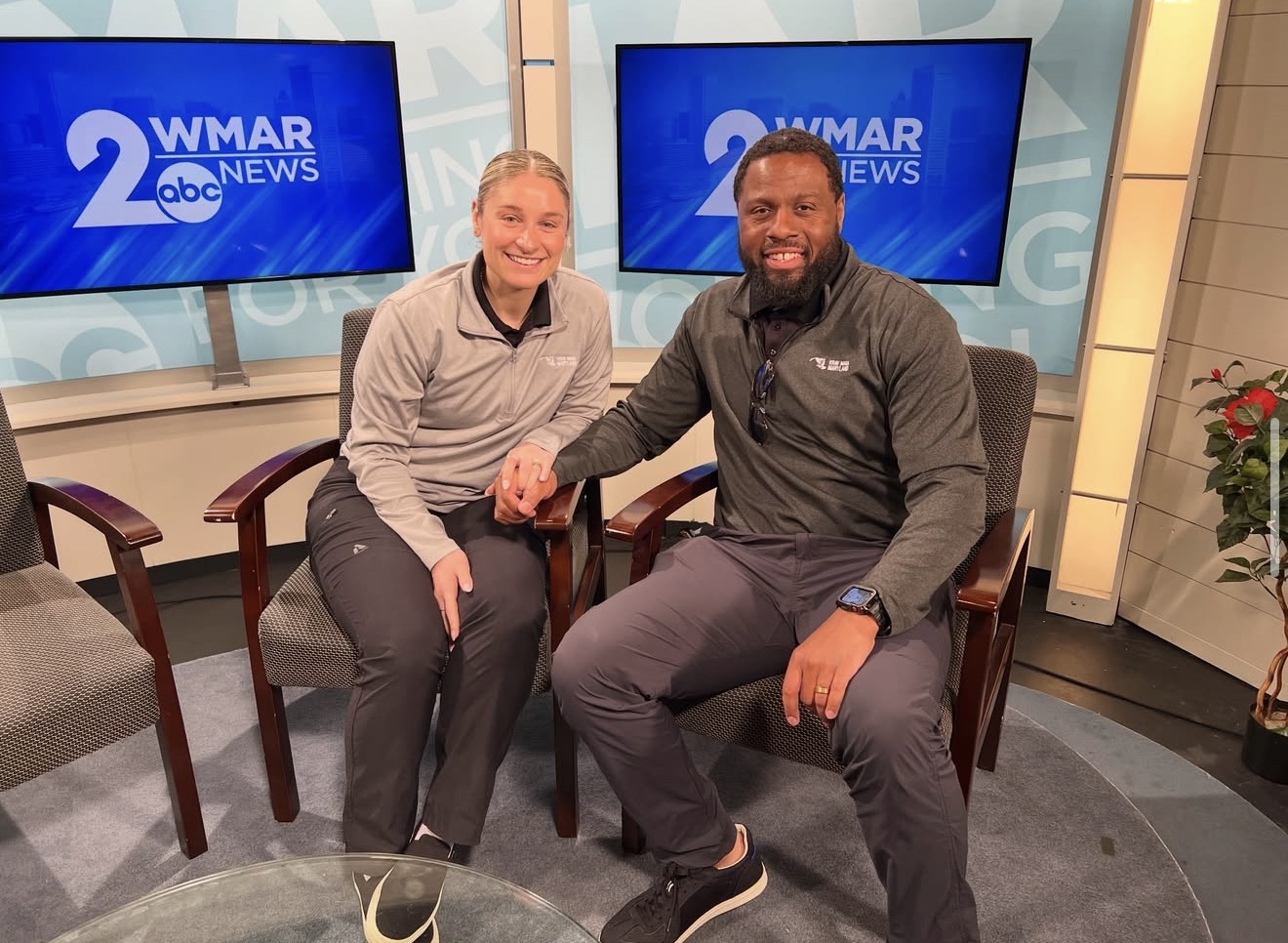Two people sitting in a TV studio, holding hands, smiling, with screens behind them displaying '2WMAR ABC NEWS'.