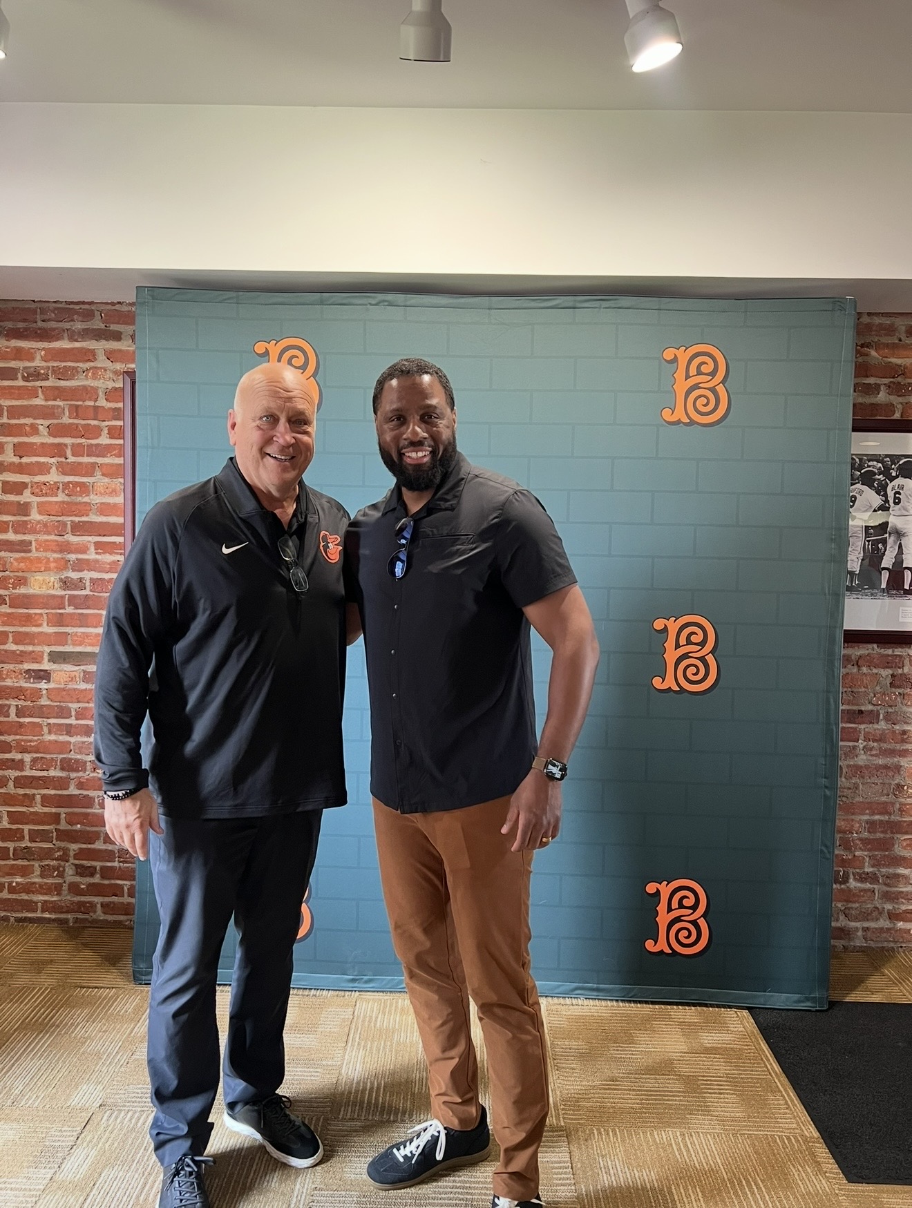 Two men stand side by side in front of a blue backdrop with orange logos, smiling indoors with brick walls and a wooden floor.