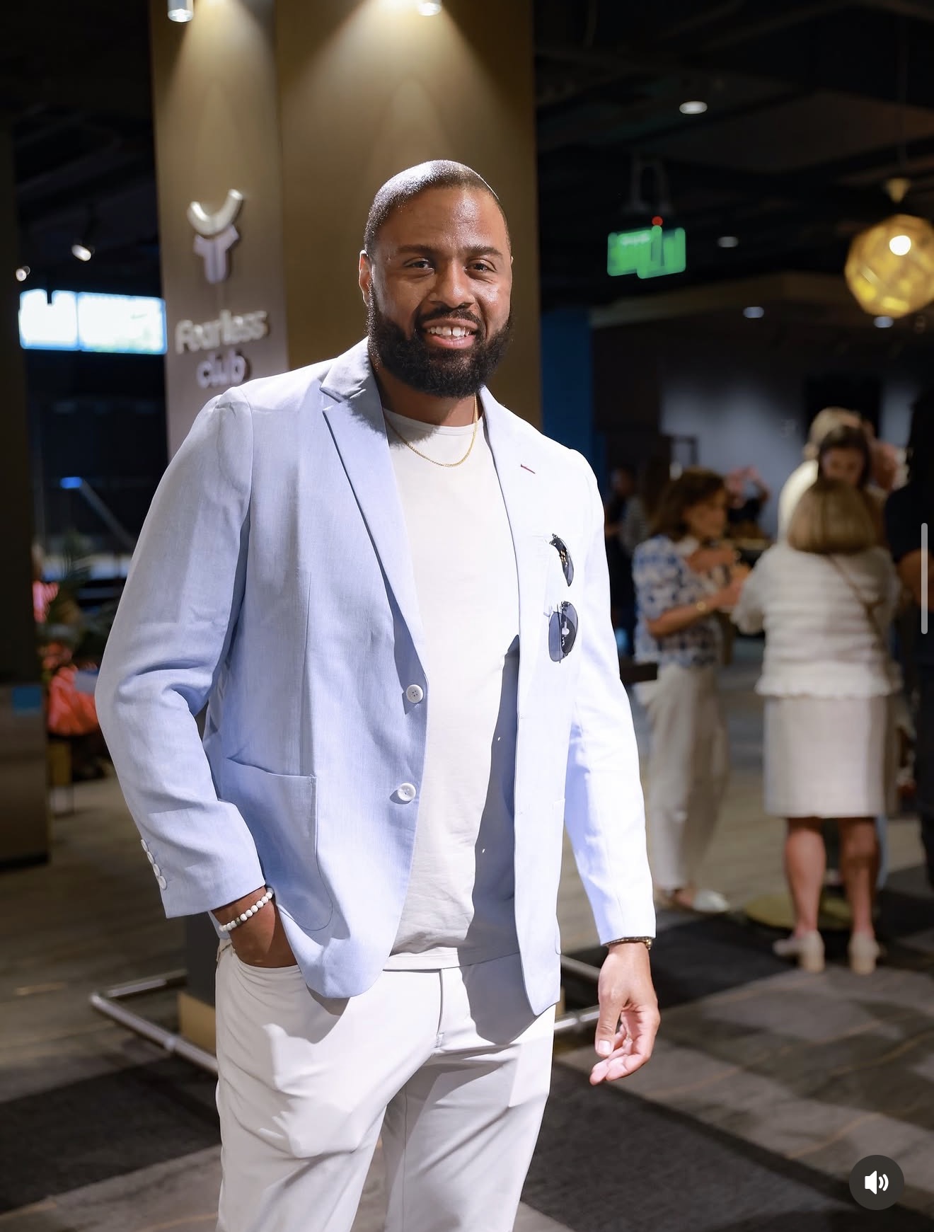 Man with beard in light blue blazer and white pants smiling at event, people in background.