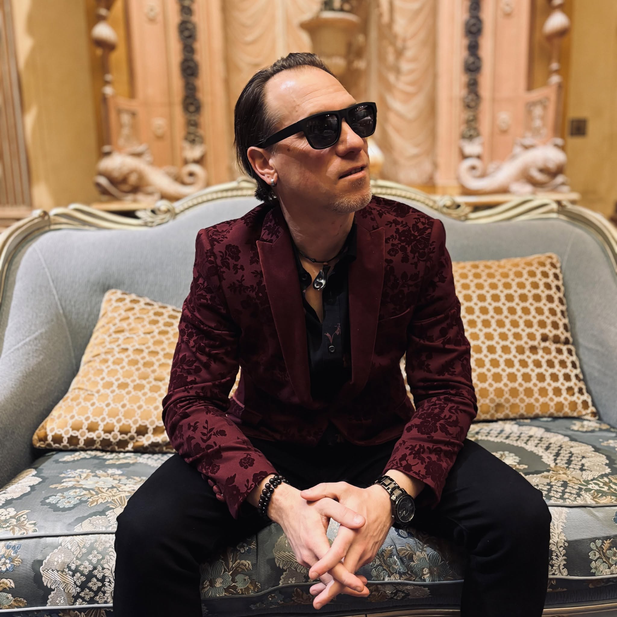 Man with dark hair, sunglasses, burgundy blazer, sitting on a sofa with patterned cushions, in a decorated room.