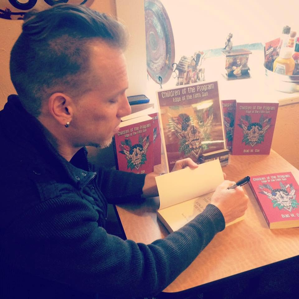 Person with short hair and earrings sitting at a table, signing a book with three books displayed upright behind them, in a well-lit room.