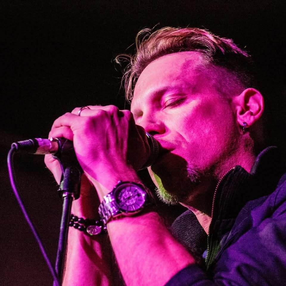 Man singing into microphone with closed eyes, wearing watch and bracelet, pink lighting, dark background.