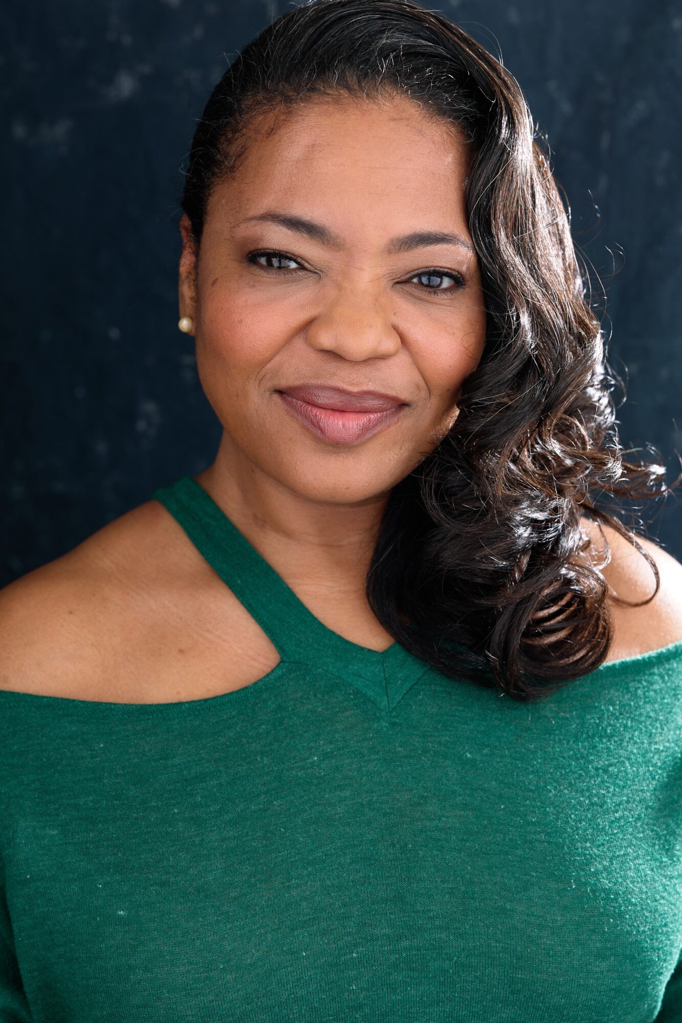 Woman with dark, wavy hair wearing a green off-shoulder top, smiling softly, dark background.