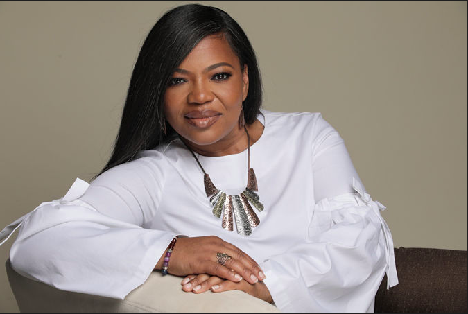 Woman with long black hair wearing a white blouse and a statement necklace, sitting with arms crossed.