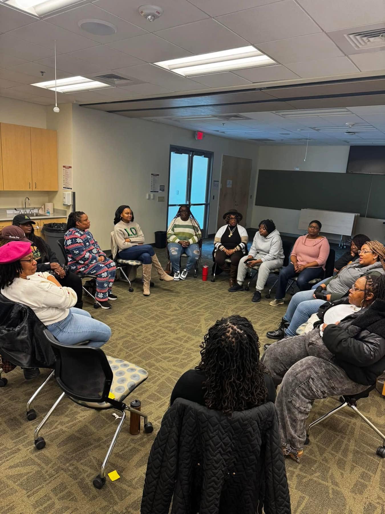 Group of people sitting in a circle in a room with a door and windows, engaged in discussion.