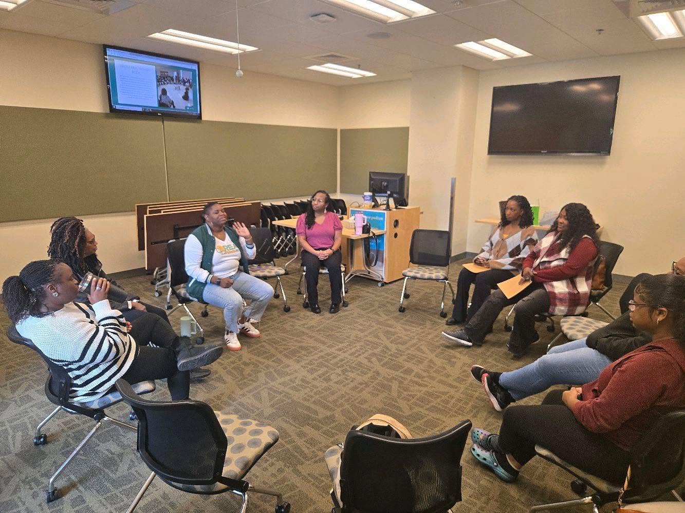 Group of people seated in a circle in a classroom or meeting room, engaging in discussion, with screens on walls and a carpeted floor.