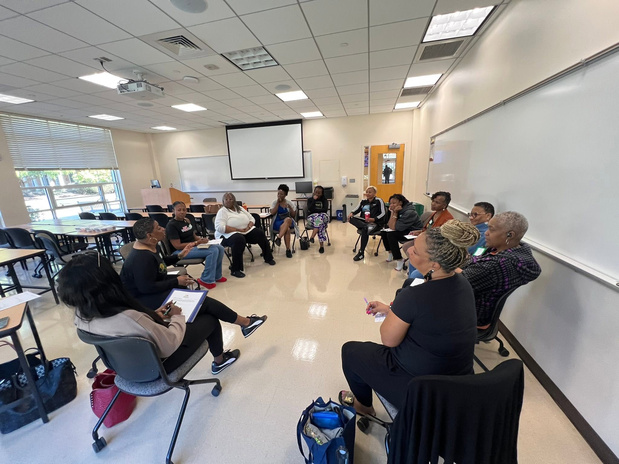 Group of people seated in a circle in a classroom, with a whiteboard and projector screen behind them.