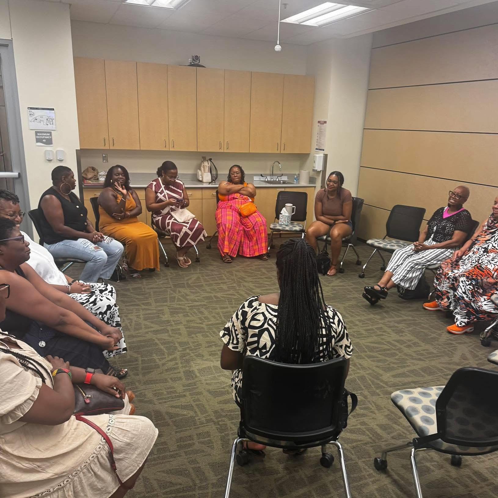 Group of women sitting in a circle in a room with beige walls and cabinets, participating in a discussion or meeting.