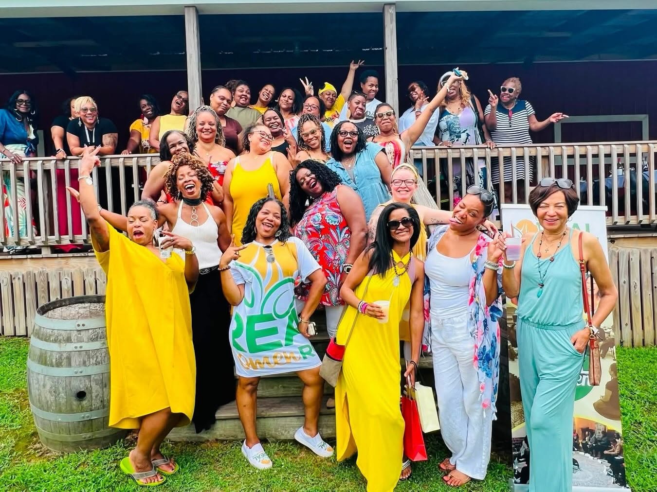 Group of diverse women smiling and posing outdoors on a balcony, some wearing bright yellow dresses, with a railing and building in background.
