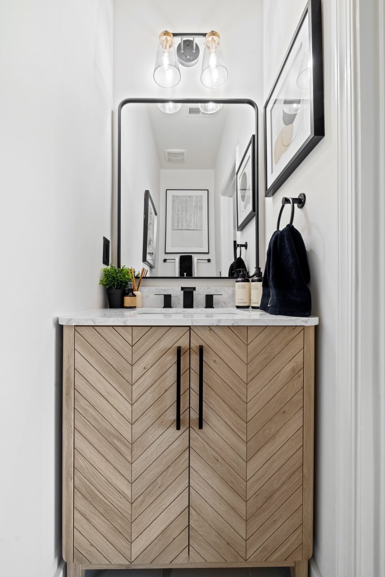 Bathroom vanity with mirror, light fixture, framed pictures, black towel, and potted plant, viewed from the doorway.