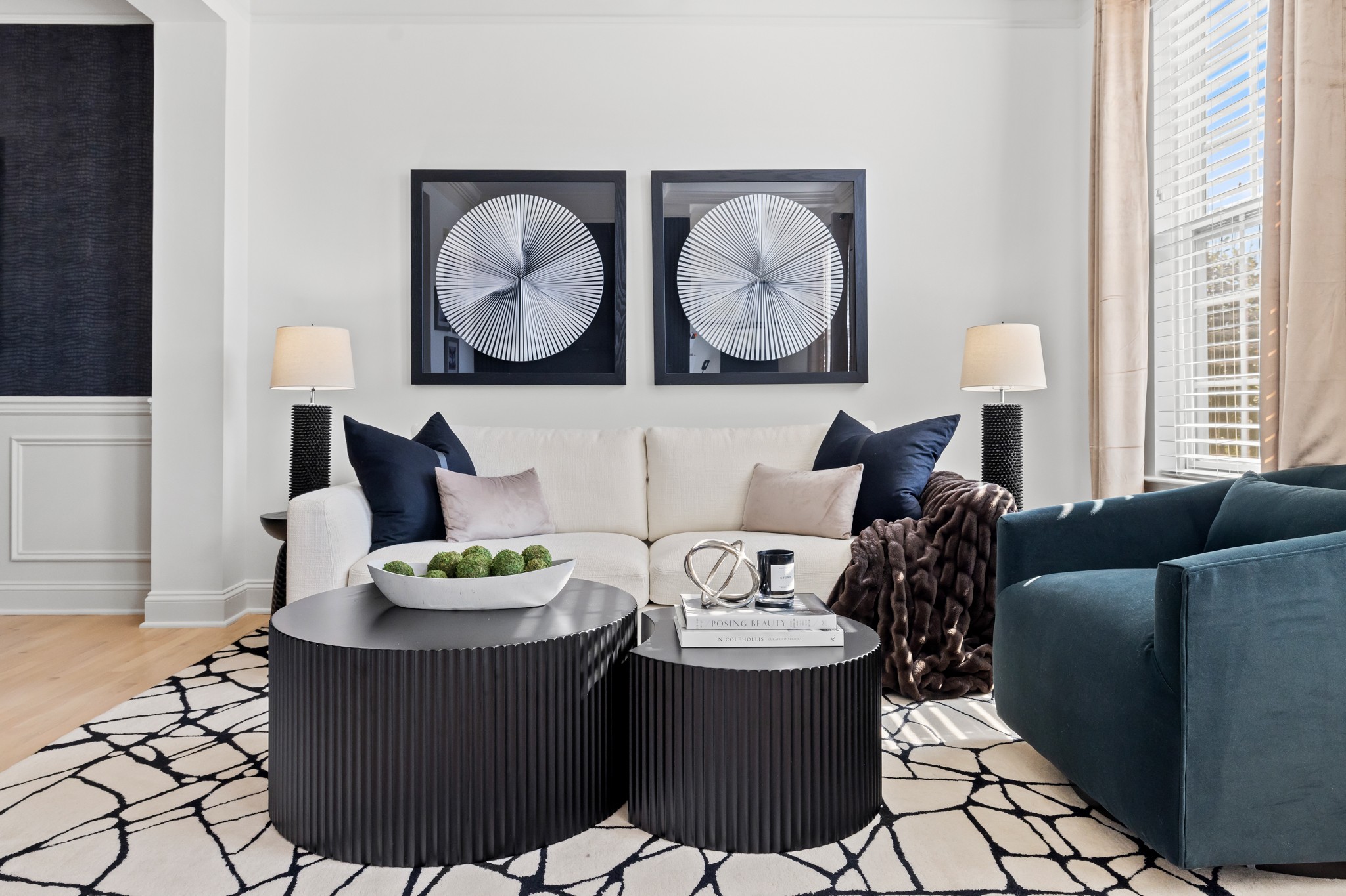 Living room with white sofa, black and blue pillows, two black coffee tables, and wall art, with natural light from windows.