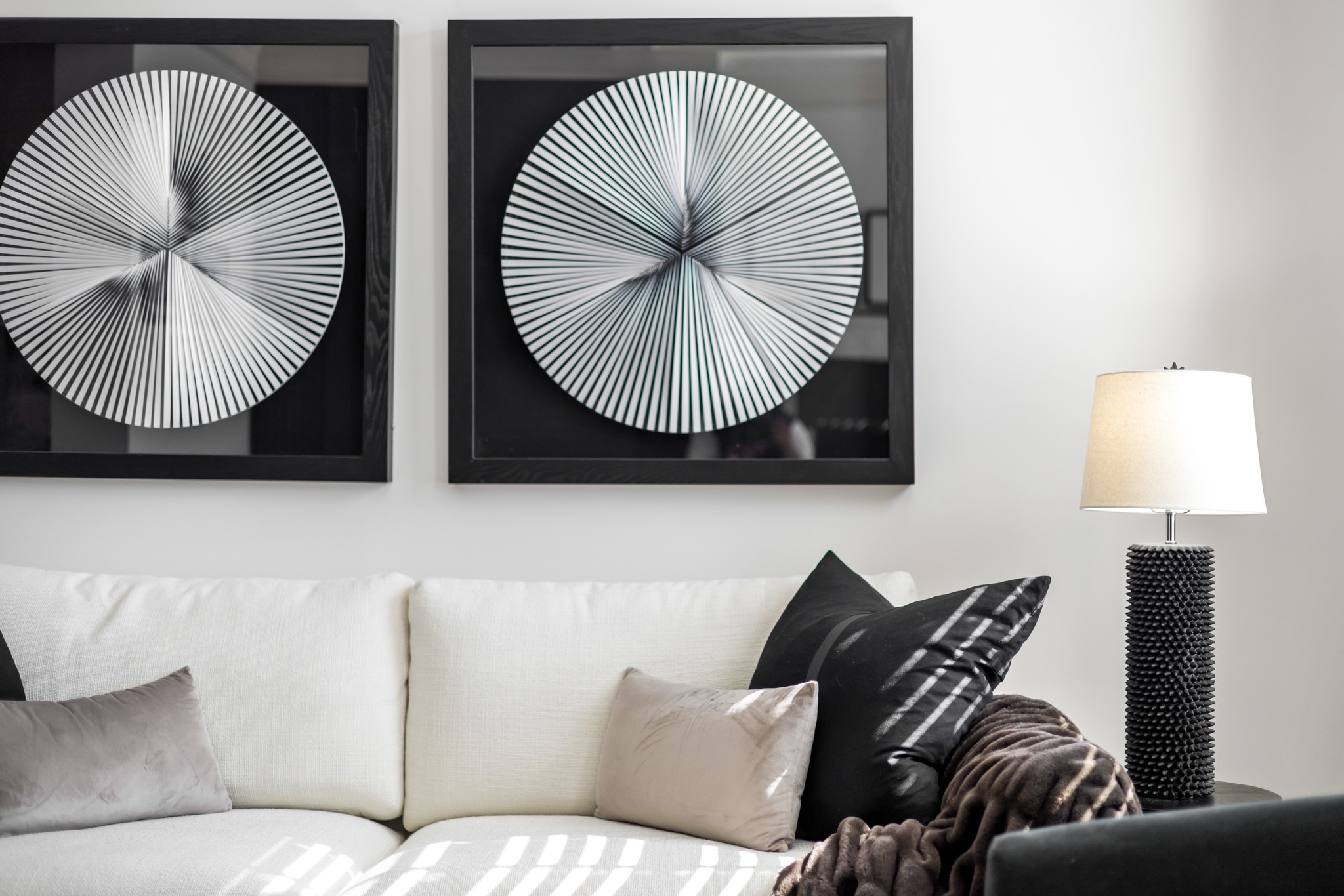 Living room with two framed circular abstract art pieces, a white sofa with pillows, and a table lamp, in a bright space.