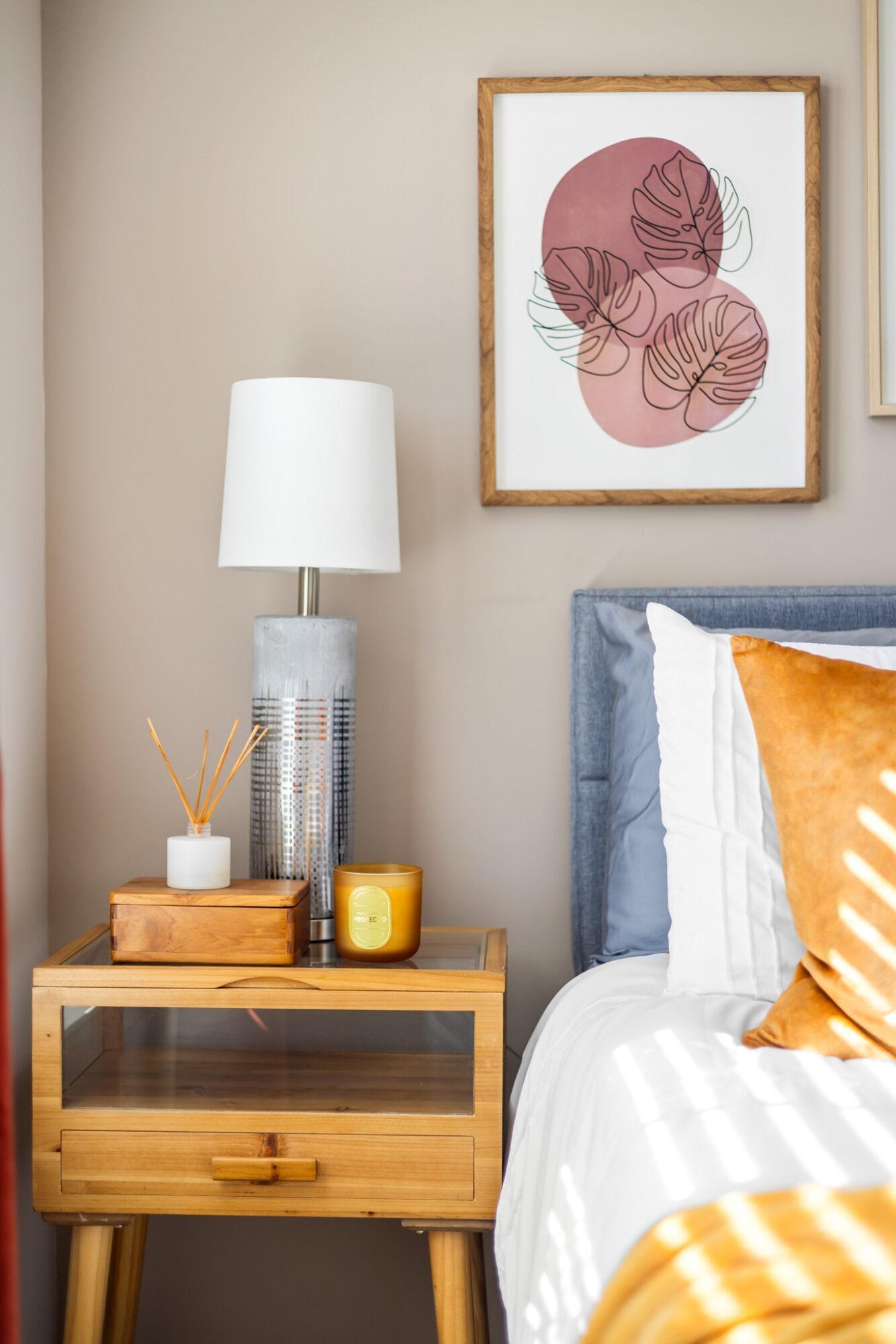 Bedroom corner with a nightstand, lamp, framed abstract art, and part of a bed with pillows.