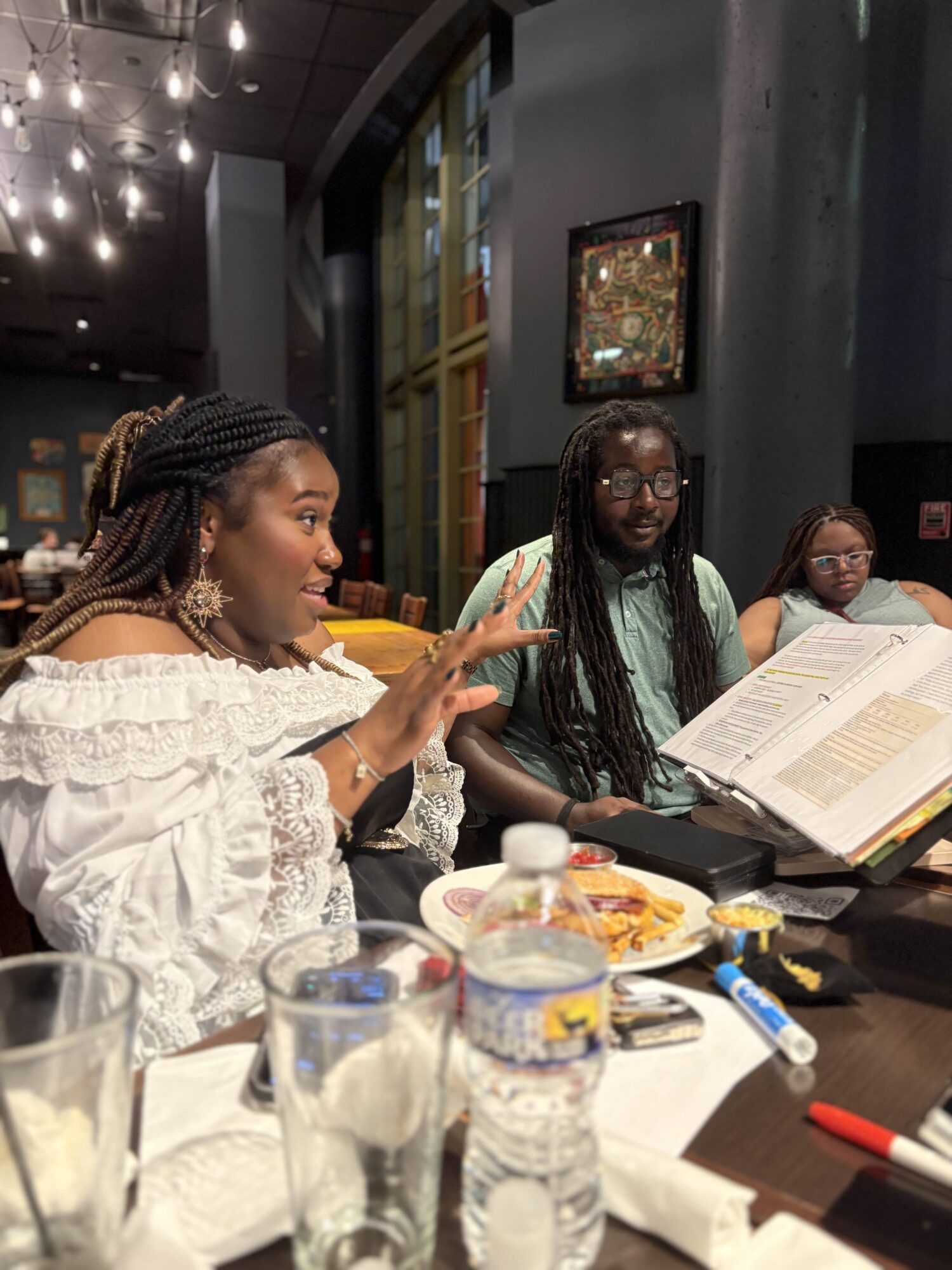 Three women sit at a table with food and drinks, engaged in conversation in a dimly lit restaurant or cafe.
