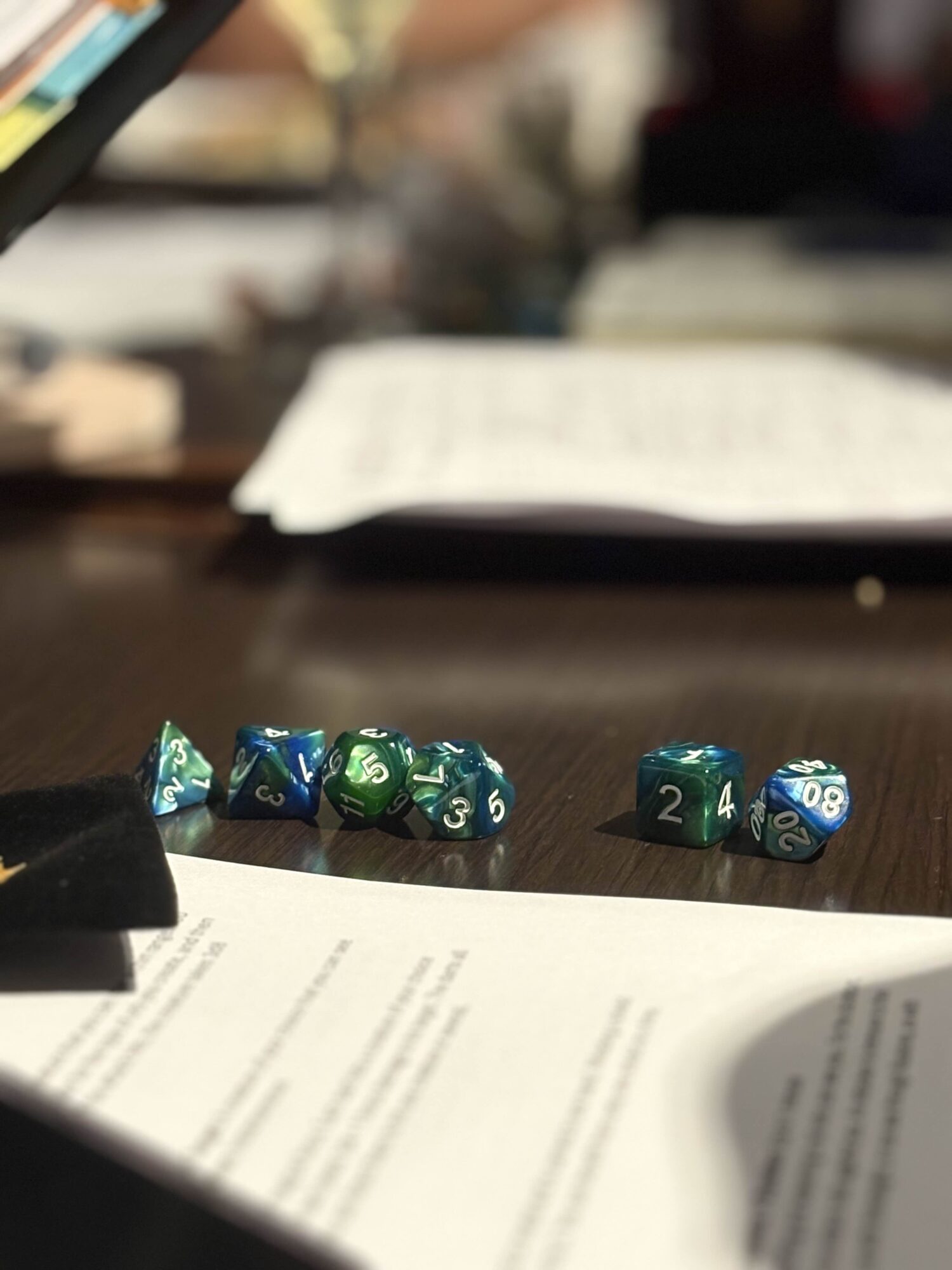 Six polyhedral dice on a table, some showing numbers, with papers and a blurred background.