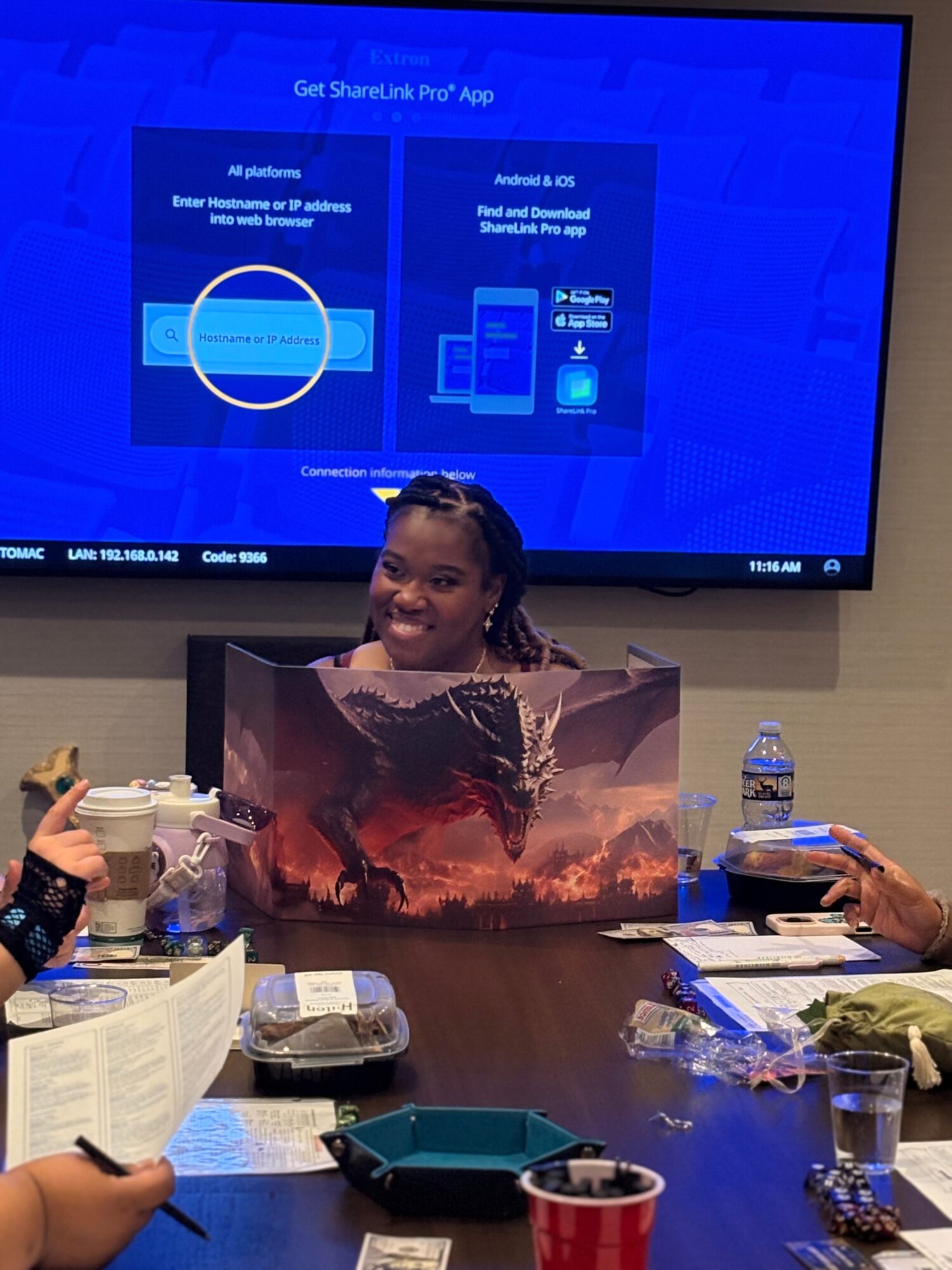 Person sitting at a table with a large fantasy-themed poster, smiling, with a TV screen behind displaying app information.