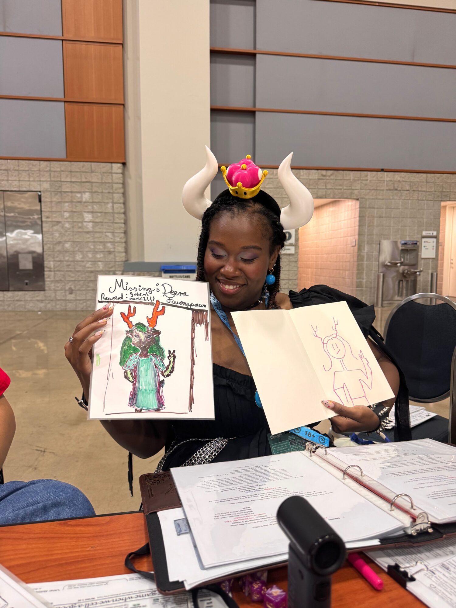 Person wearing a unicorn headband holding a drawing of a woman in a green dress, sitting at a table with art supplies.