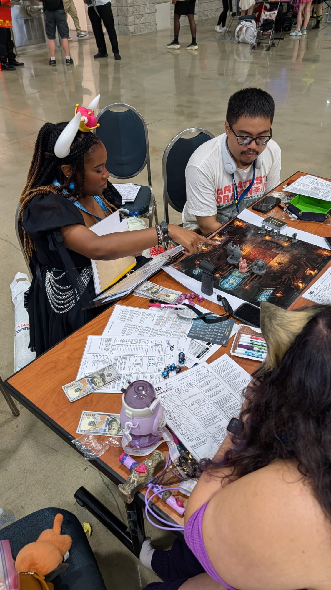 A young girl with a unicorn headband and a man with glasses sit at a table playing a tabletop game, surrounded by papers and game pieces.