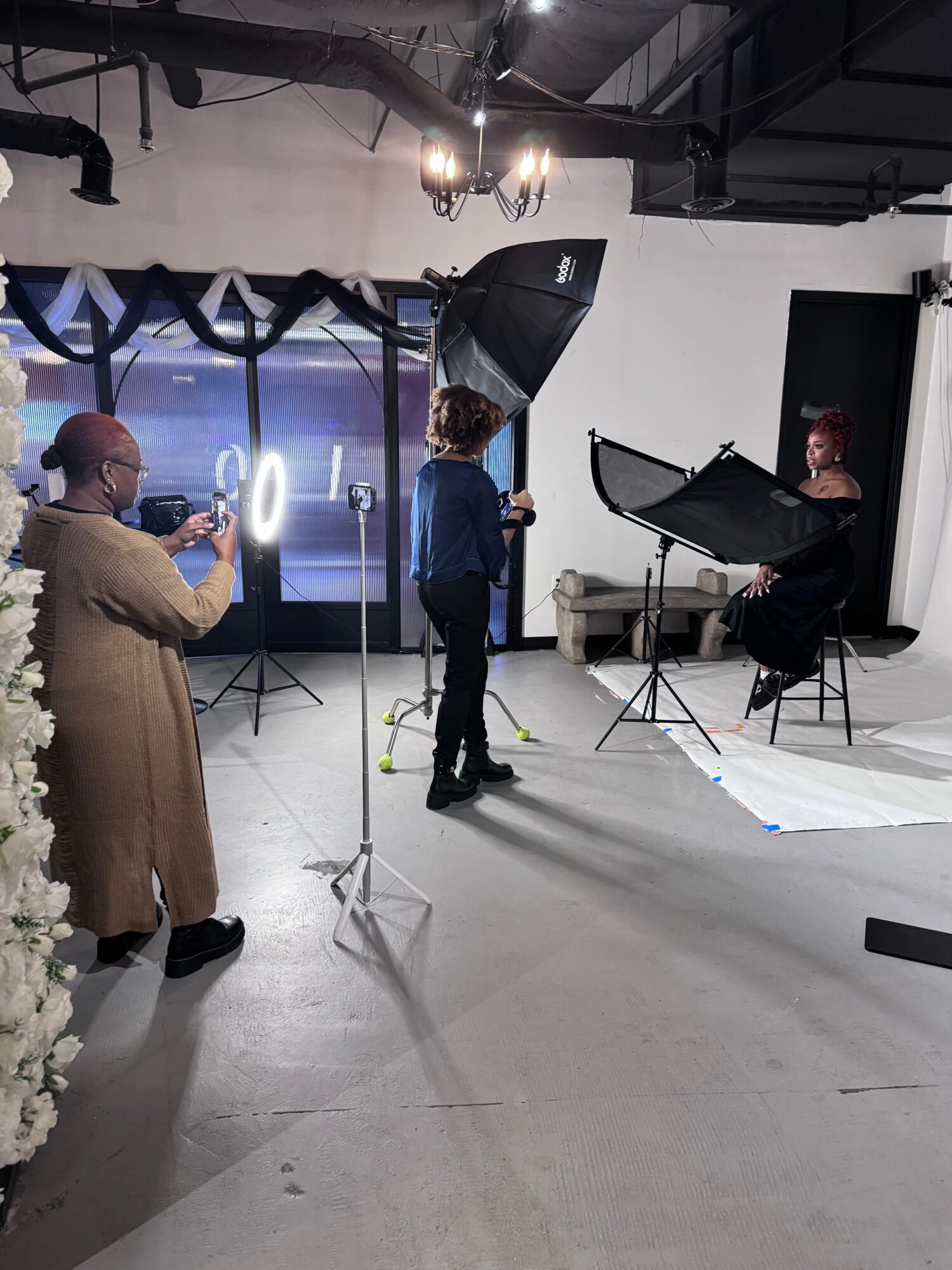People in a photography studio with lighting and a backdrop, capturing a seated person with a large umbrella light.