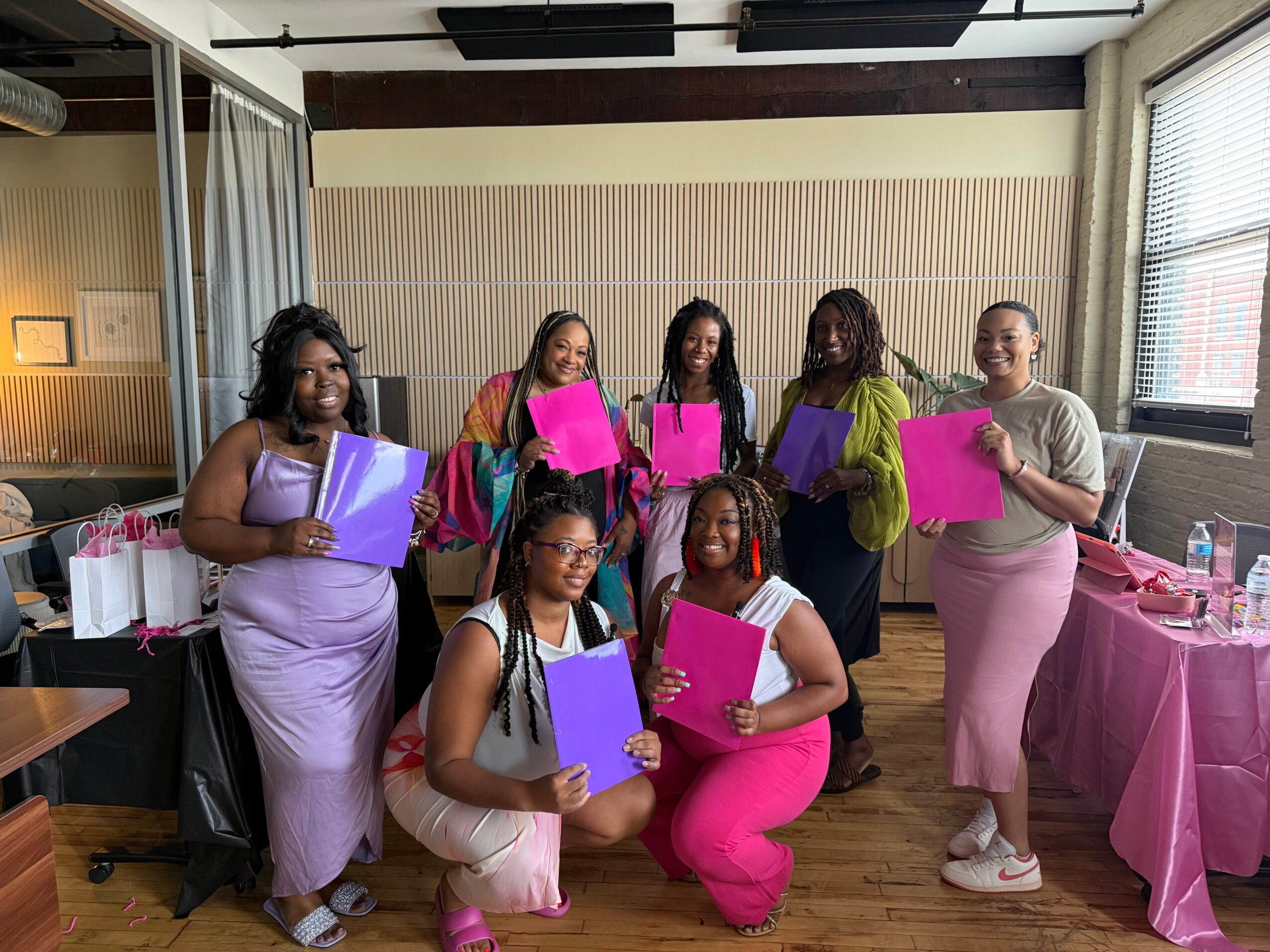 Seven women holding pink and purple folders, smiling indoors with a table of gifts and decorations nearby.