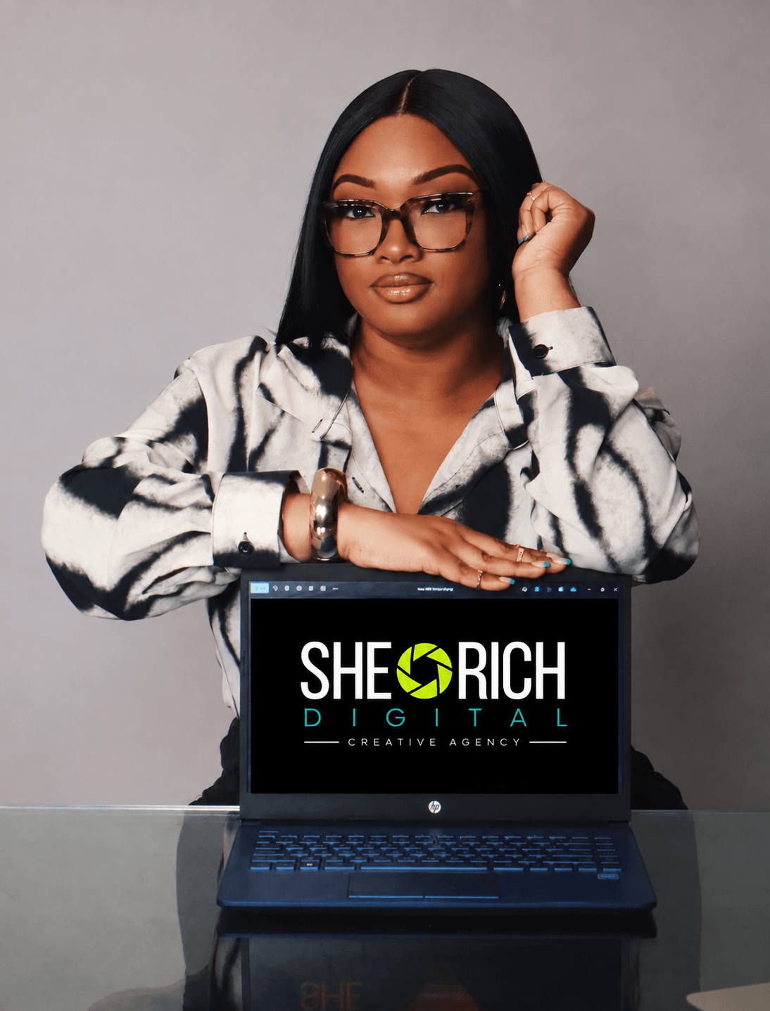 Woman with glasses and long black hair, wearing a patterned shirt, resting her chin on her hand, with a laptop displaying 'SHE RICH DIGITAL' in front of her.