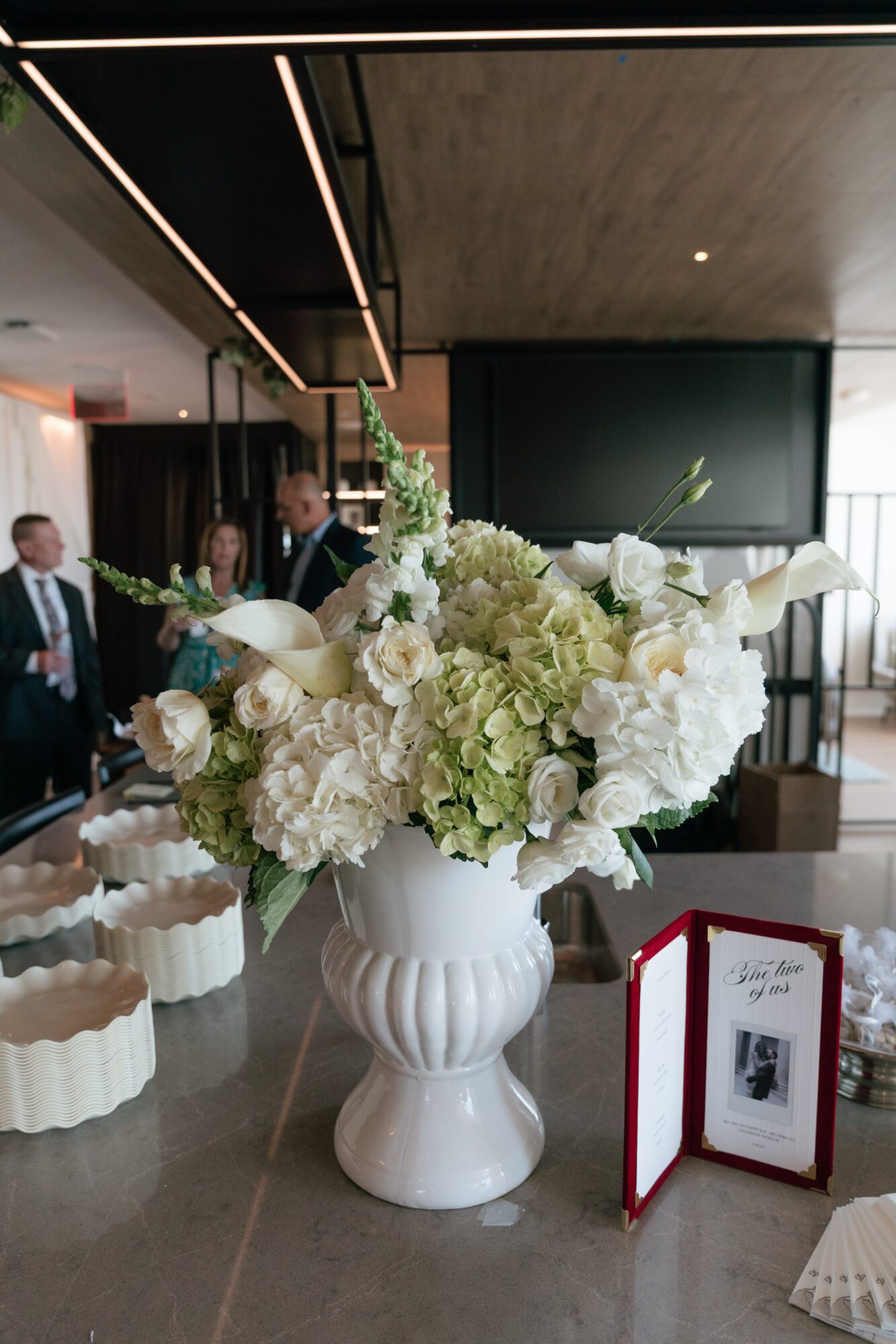 White floral arrangement in a vase on a table with people in background, modern interior setting.