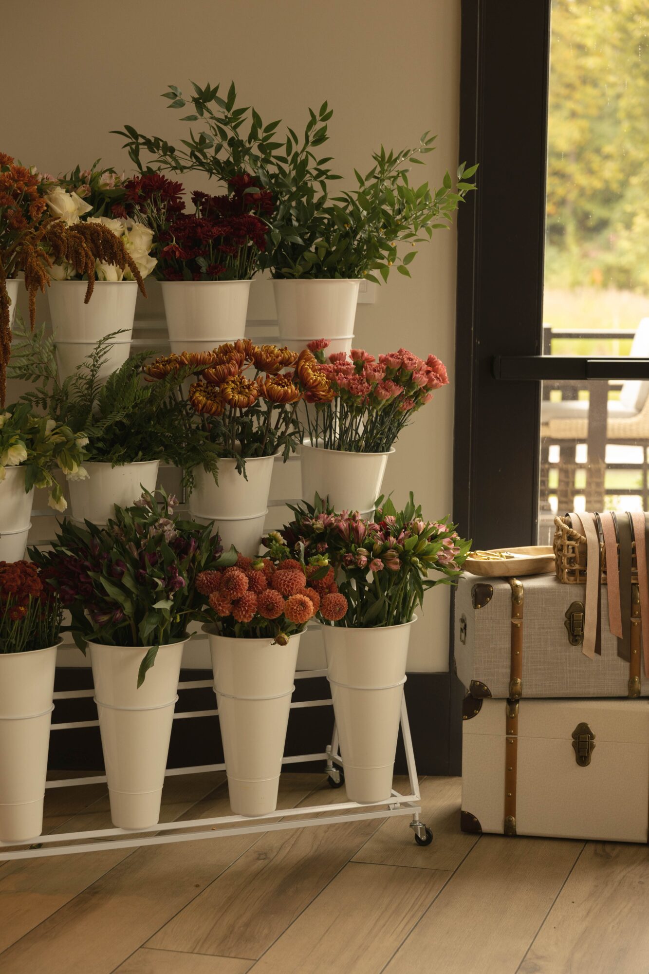 Indoor plant display with flowers in white pots near a window, with a suitcase beside them, wooden floor visible.