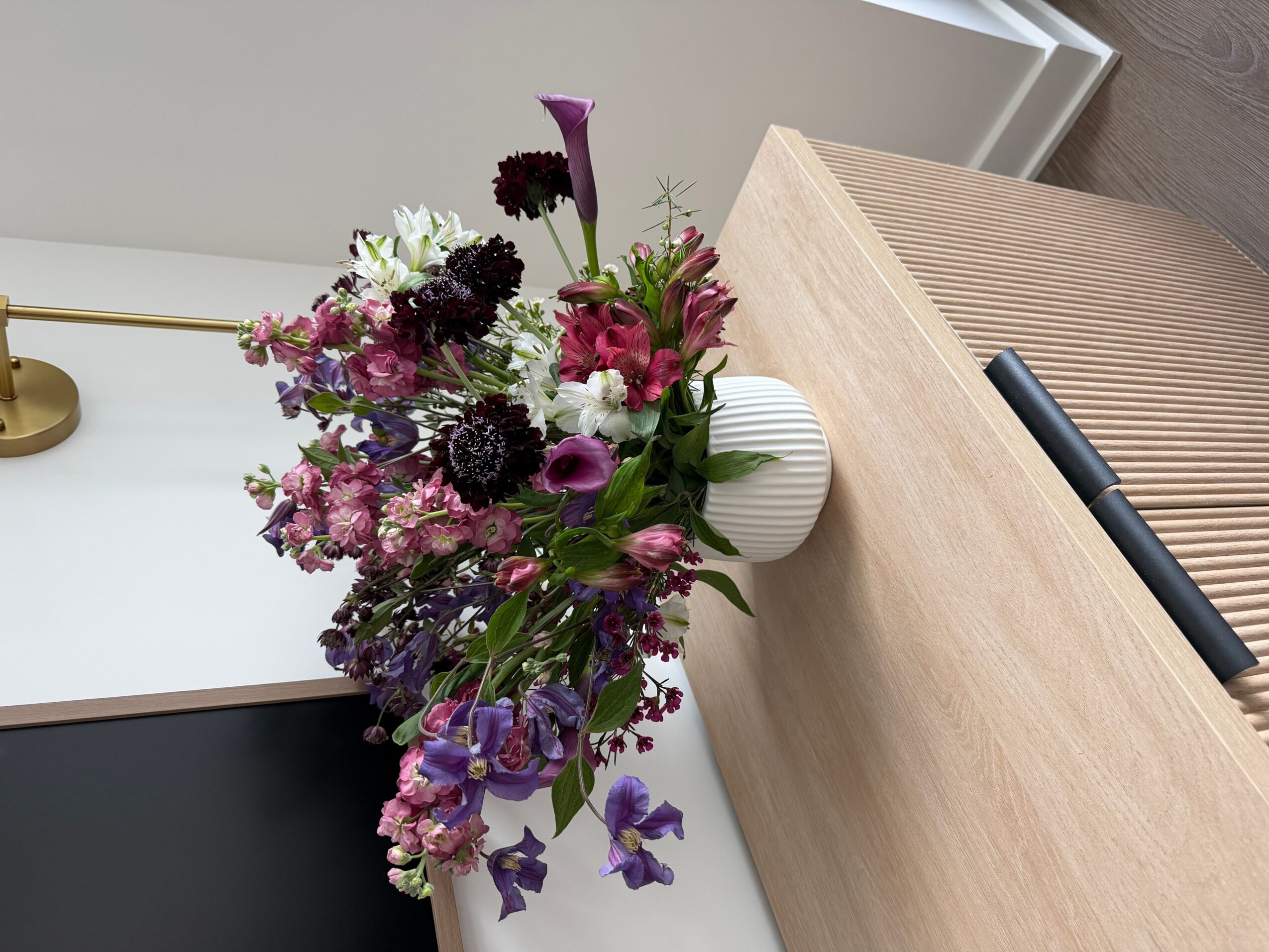 Colorful flower arrangement in a white vase on a wooden table, with a white wall and a lamp in the background.