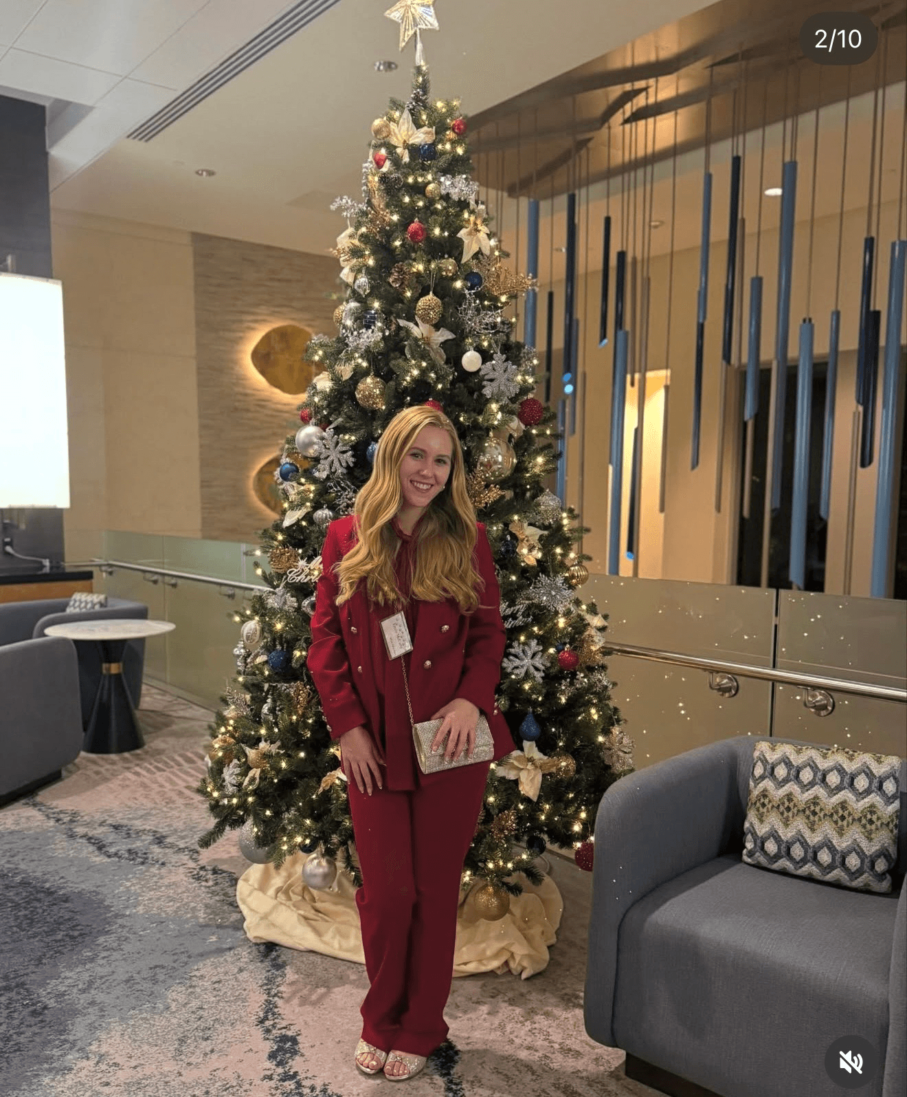 Woman in red outfit standing in front of decorated Christmas tree indoors, holding a gift bag, smiling.