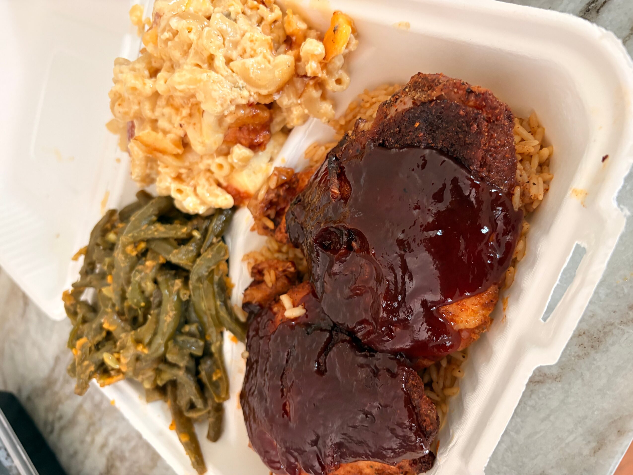 Meal with mashed potatoes, green beans, and a meatloaf with dark sauce in a white container.