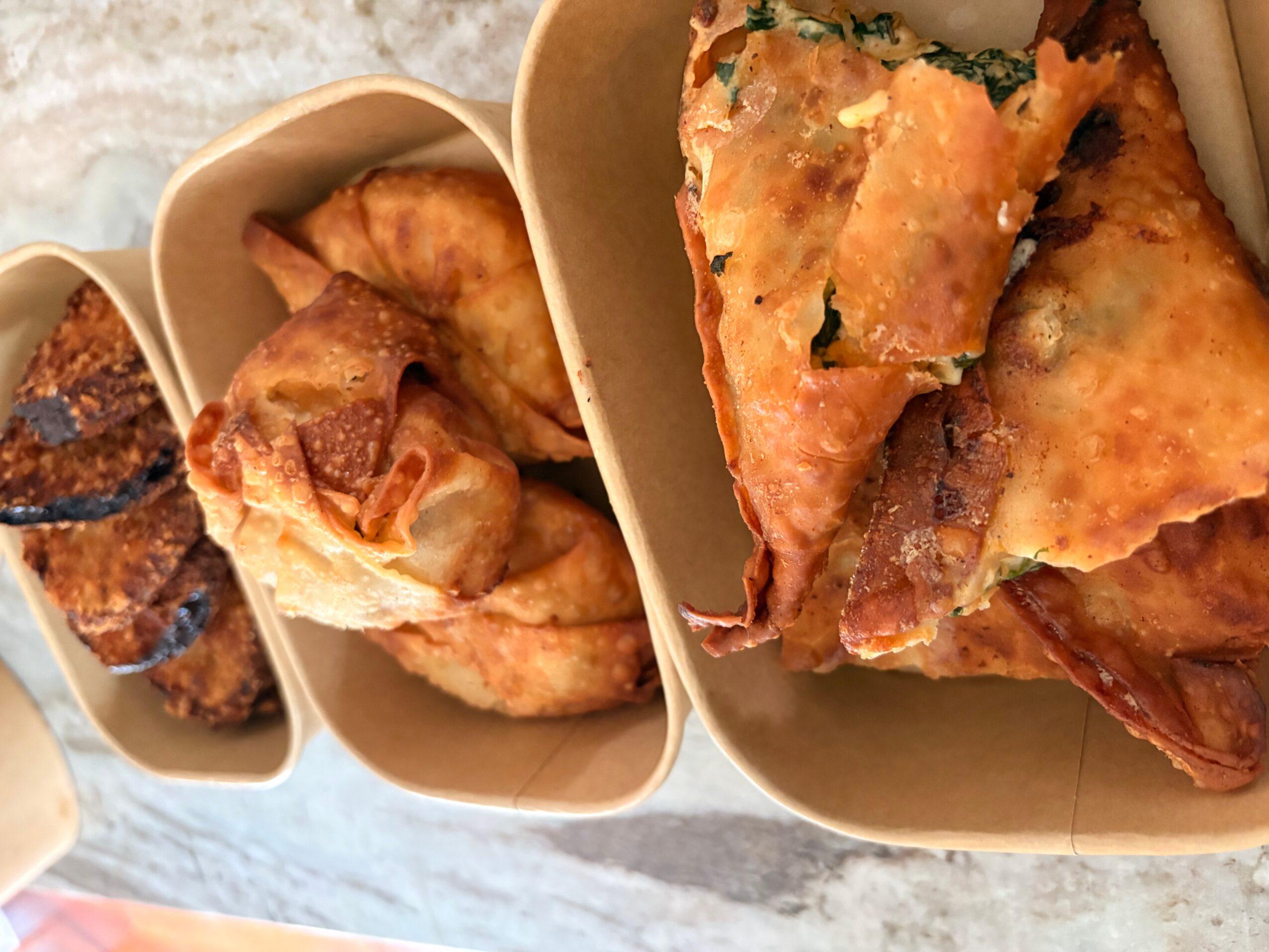 Three food items in paper trays: fried chicken, spring rolls, and a slice of pizza with cheese and toppings.