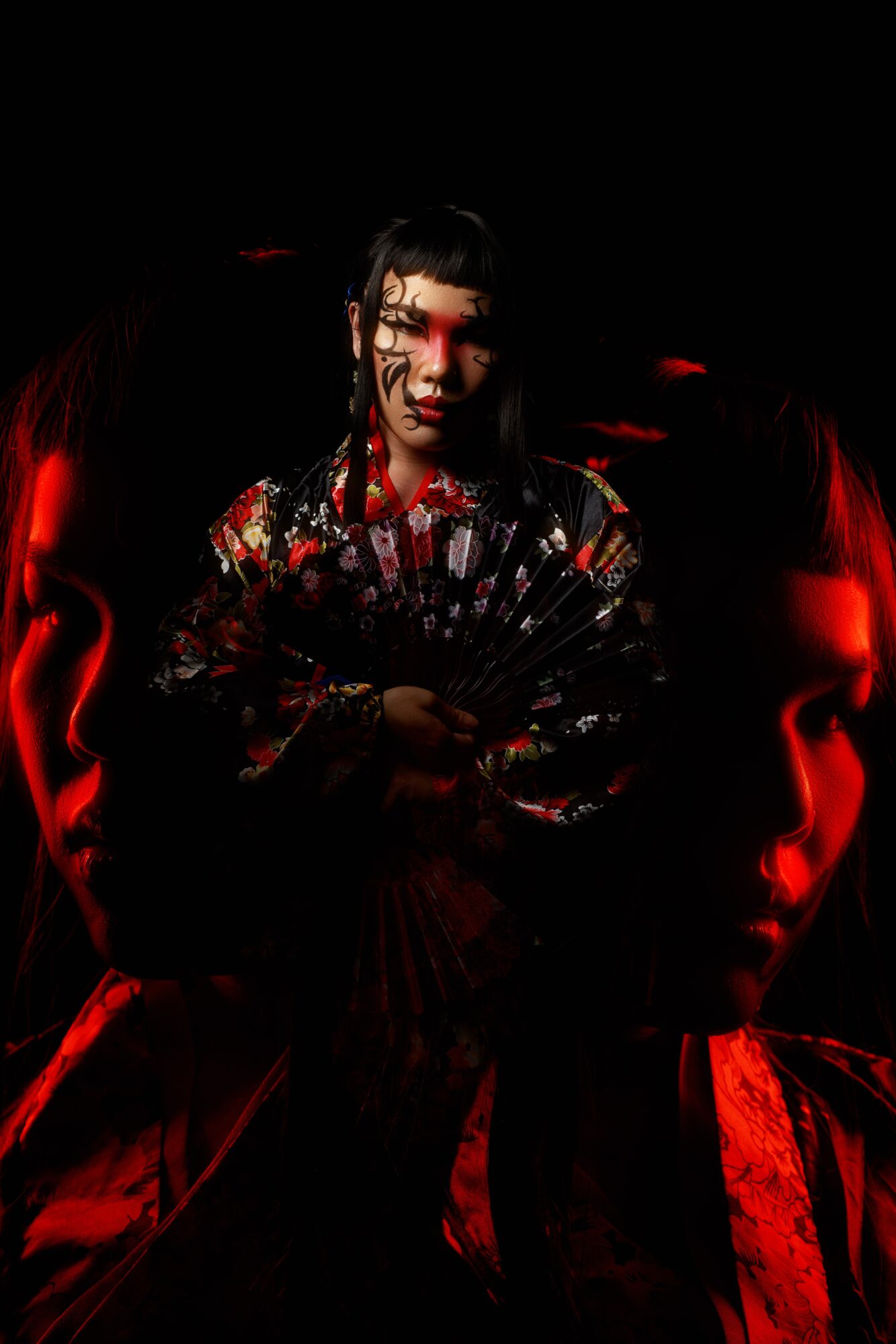 Person in traditional clothing holding a fan, with dark background and red lighting, face partially shadowed.