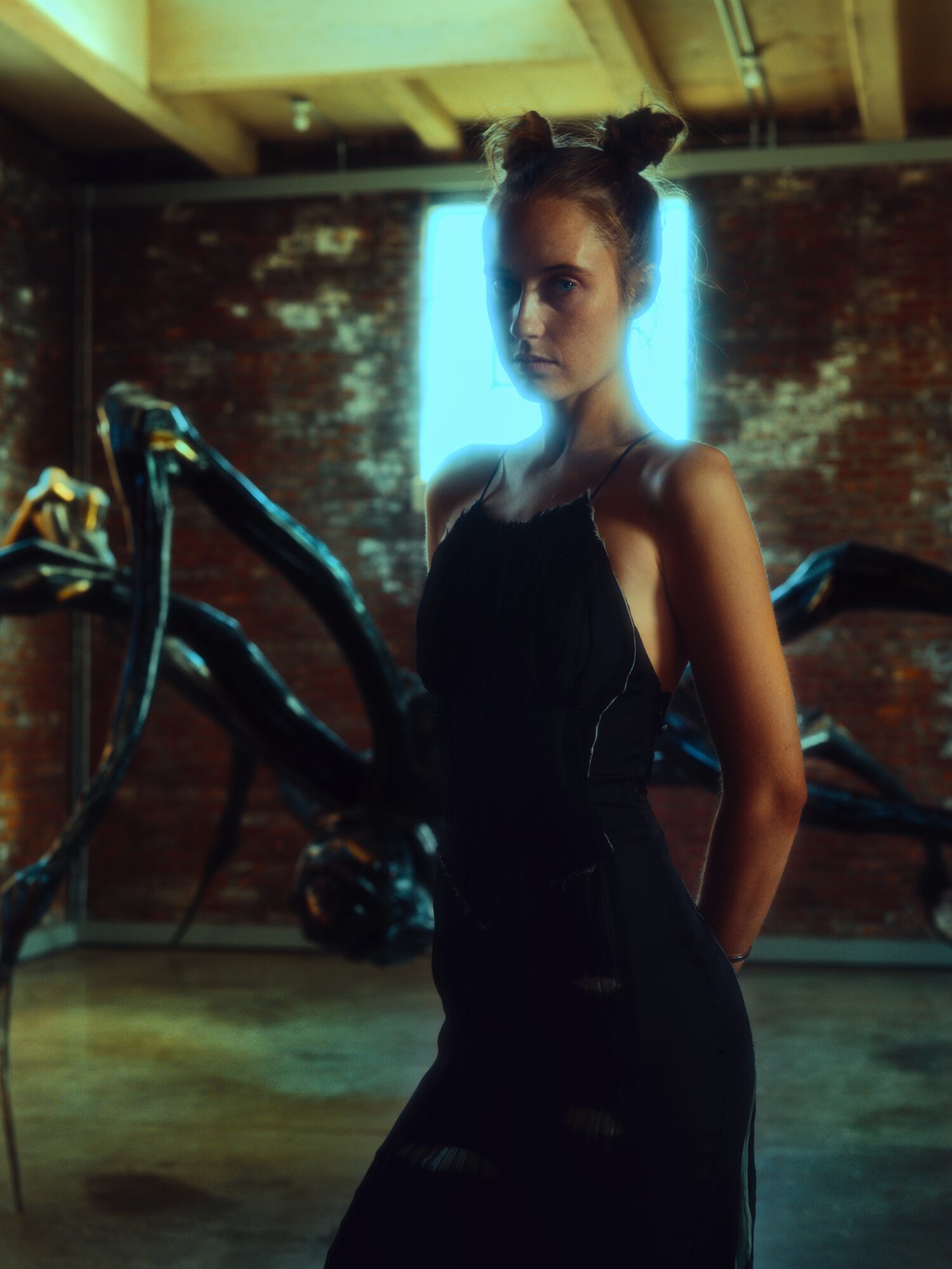 Young woman with two buns hairstyle in a dark dress, standing in a dimly lit room with a bright light behind her.