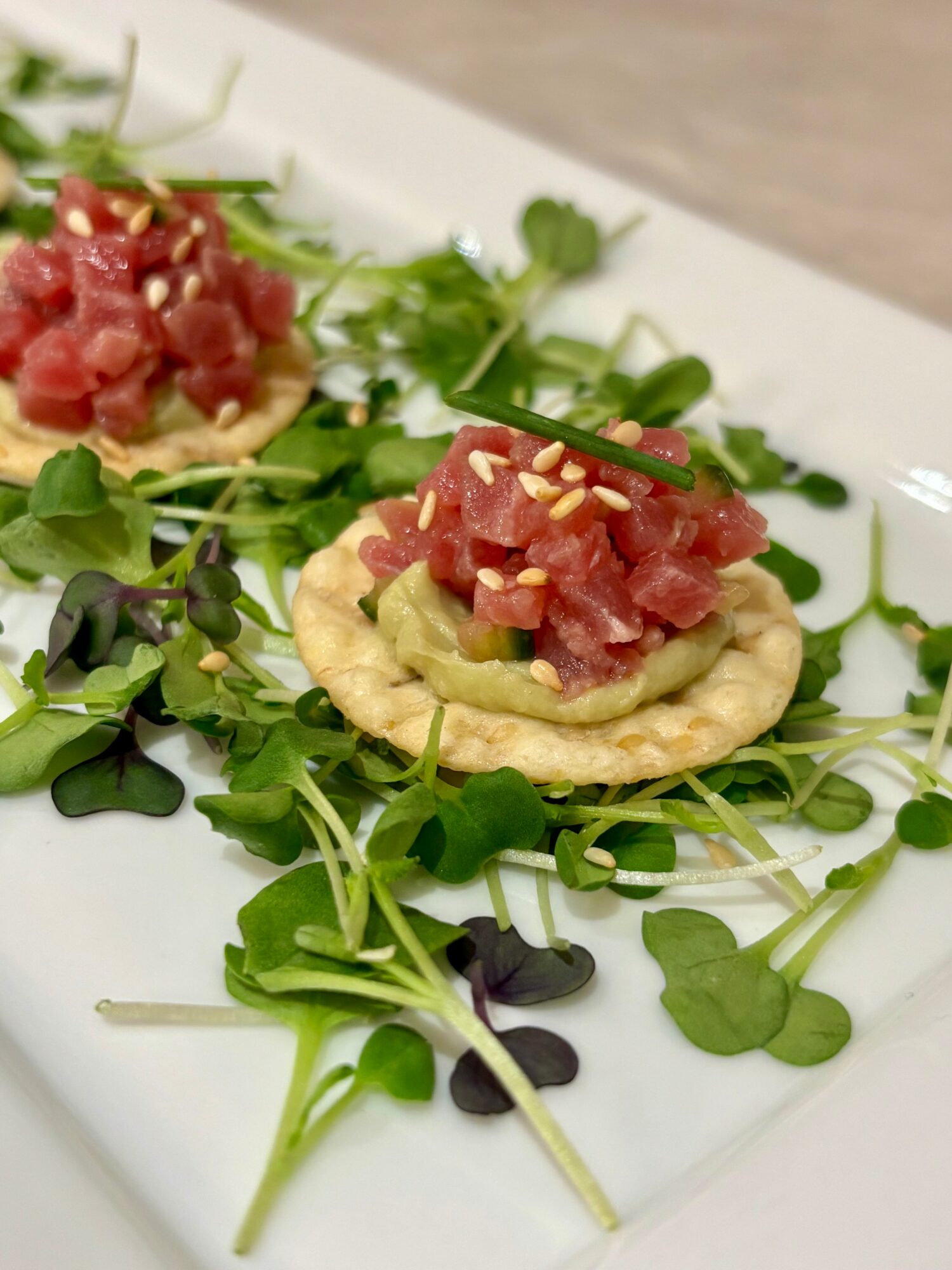 Two small appetizers with diced raw fish on crispy bases, garnished with microgreens and chopped nuts, on a white plate.