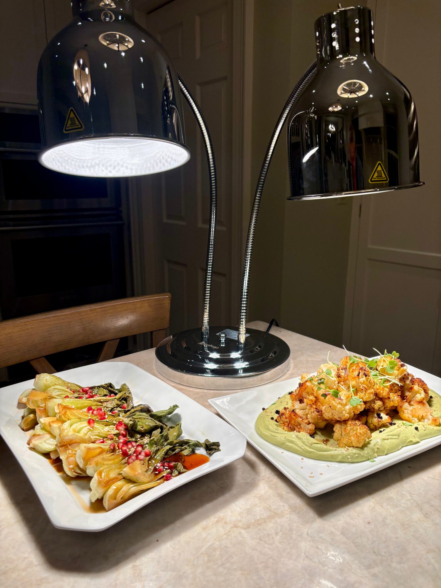 Two plates of food on a table with a dual-armed lamp overhead, one plate with pasta and the other with fried food.