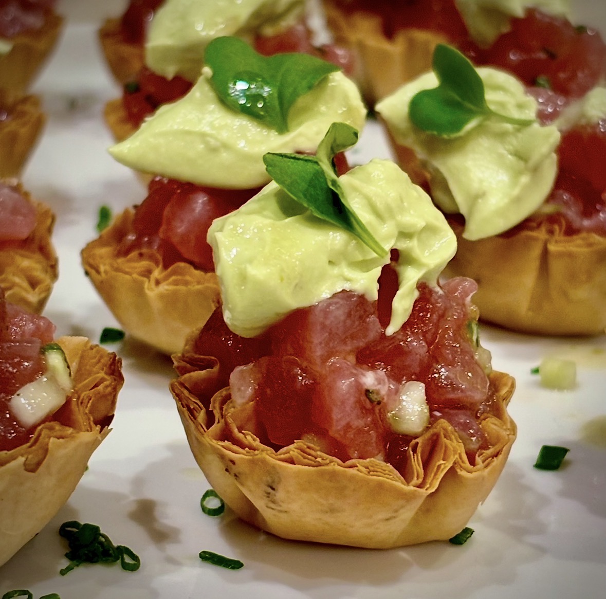 Mini tartlets filled with diced meat, topped with avocado and small green herbs, on a white surface.
