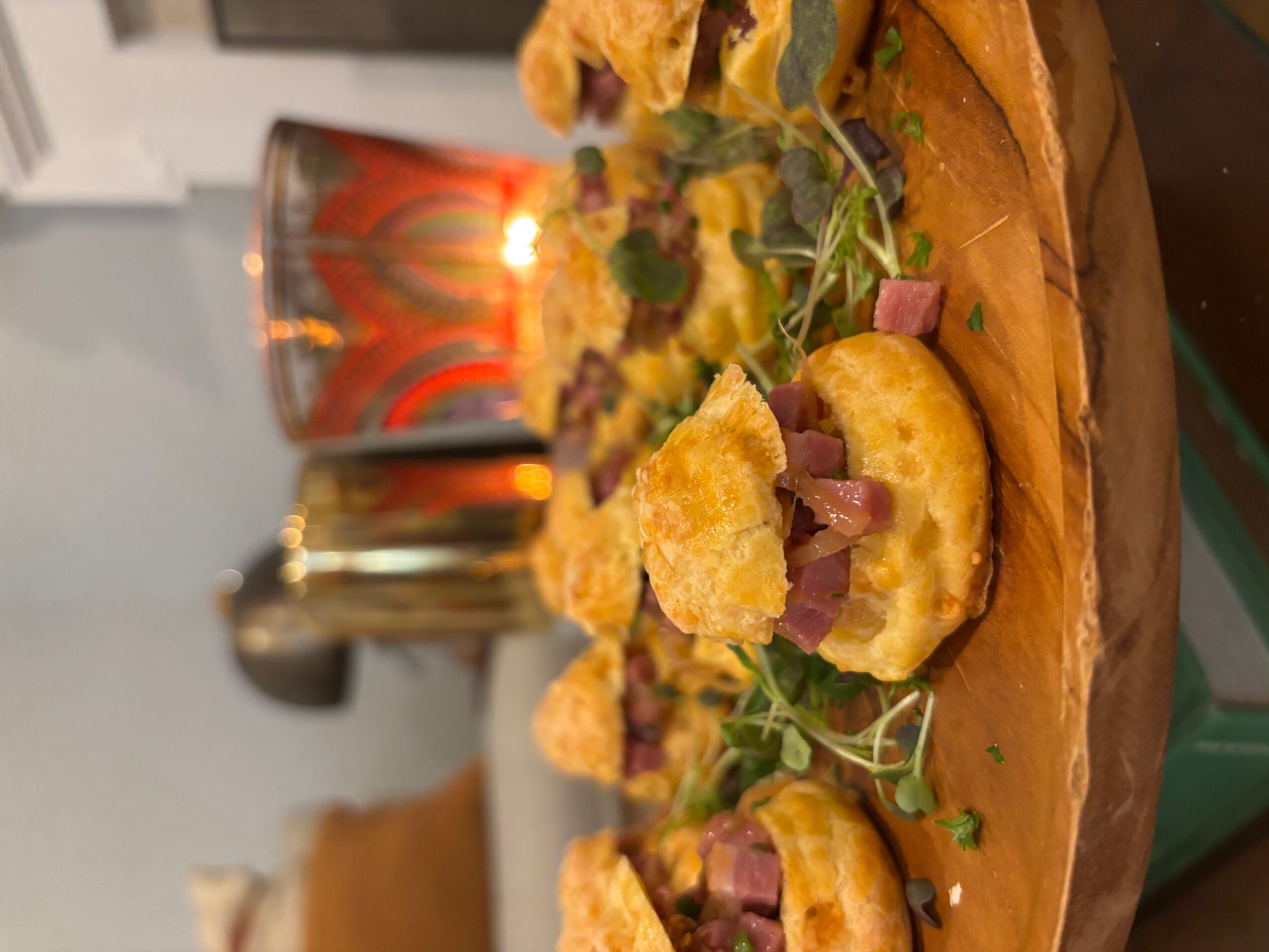 Close-up of a pizza with toppings including cheese, ham, and greens on a wooden board, with a drink in the background.