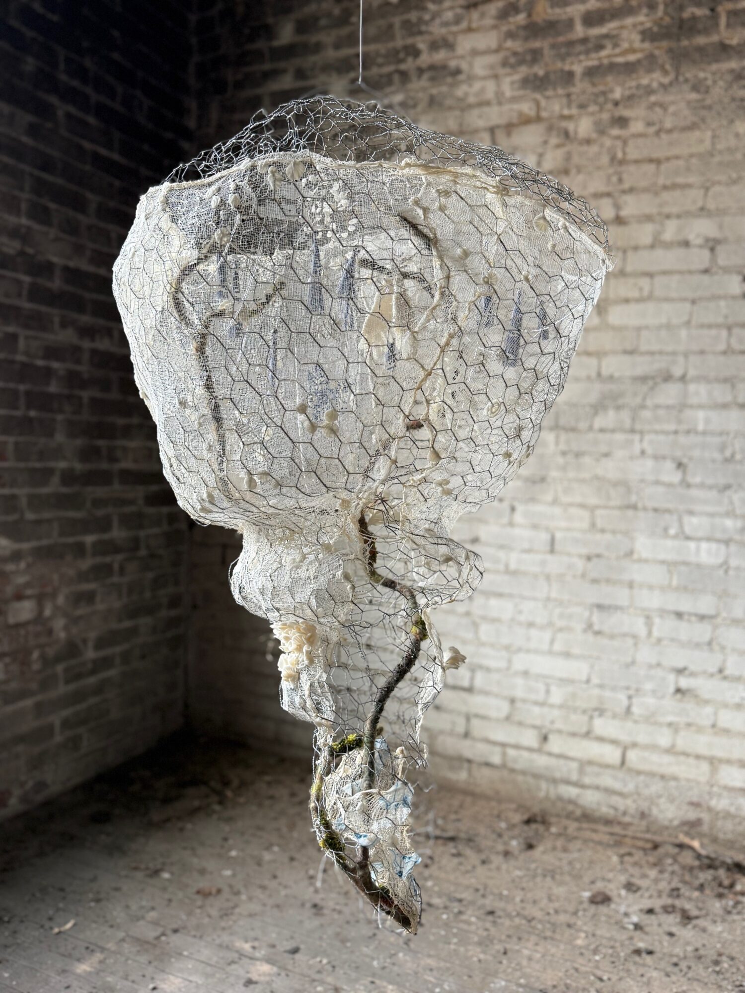 Sculpture of a face with wire mesh and stone, mounted on a stand against a brick wall, in an indoor setting.