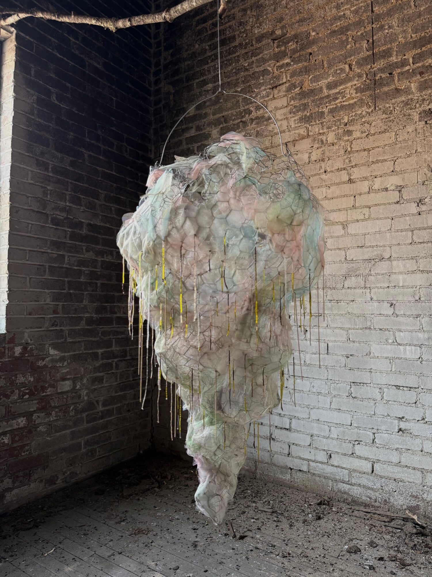 Hanging abstract sculpture made of layered, colorful, textured materials in a corner with brick walls.