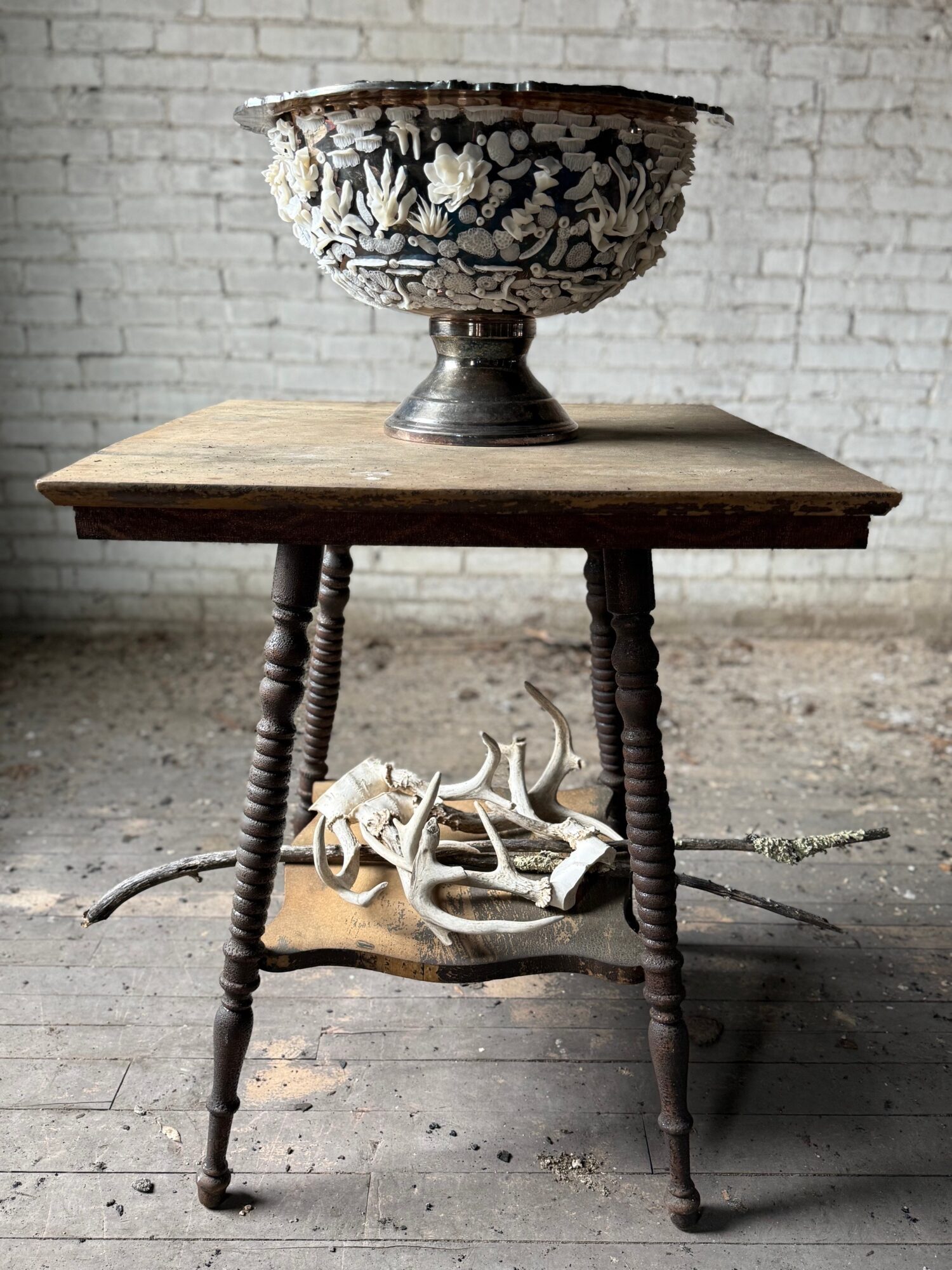 Wooden table with a decorative bowl on top, and animal skulls on a lower shelf, against a brick wall background.